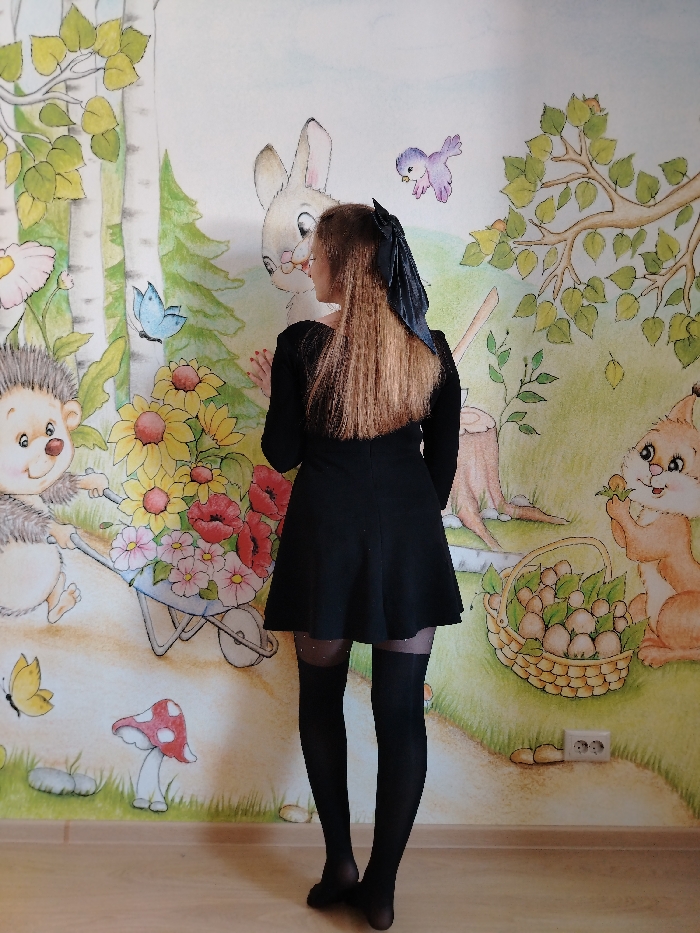 An interior scene in the city of Chaikovsky, Russia, displays a person facing a wall mural. The individual, appearing female, is positioned with her back predominantly to the viewer, her head turned to her left, observing the mural. She possesses light brown hair tied with a dark ribbon and wears a long-sleeved black dress, black sheer over-the-knee tights featuring a glittered band above the knee, and black foot coverings. Her right hand is in contact with the mural. The mural itself is a colorful, cartoon-style depiction of a forest or garden environment, featuring anthropomorphic animals: a hedgehog pushing a wheelbarrow full of flowers, a rabbit, a flying purple bird, and a squirrel holding an orange object next to a basket filled with mushrooms. Additional elements include birch trees, evergreen trees, various flowers (sunflowers, poppies, daisies), a blue butterfly, a yellow butterfly, and a red-capped mushroom. A white double electrical outlet is present on the lower right section of the wall. The floor is constructed of light-toned wood.