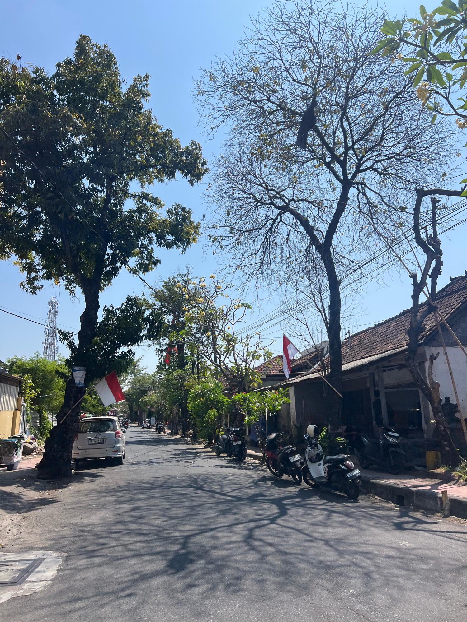 The image shows a street in Denpasar, Indonesia, on a sunny day with a clear blue sky. A white car is parked on the left side of the street, with an Indonesian flag attached to a tree in front of it. Several motorcycles are parked along the right side of the street. The street is lined with houses and trees. A cell tower is visible in the distance on the left side of the image. The scene appears to be a typical residential street in a tropical climate.