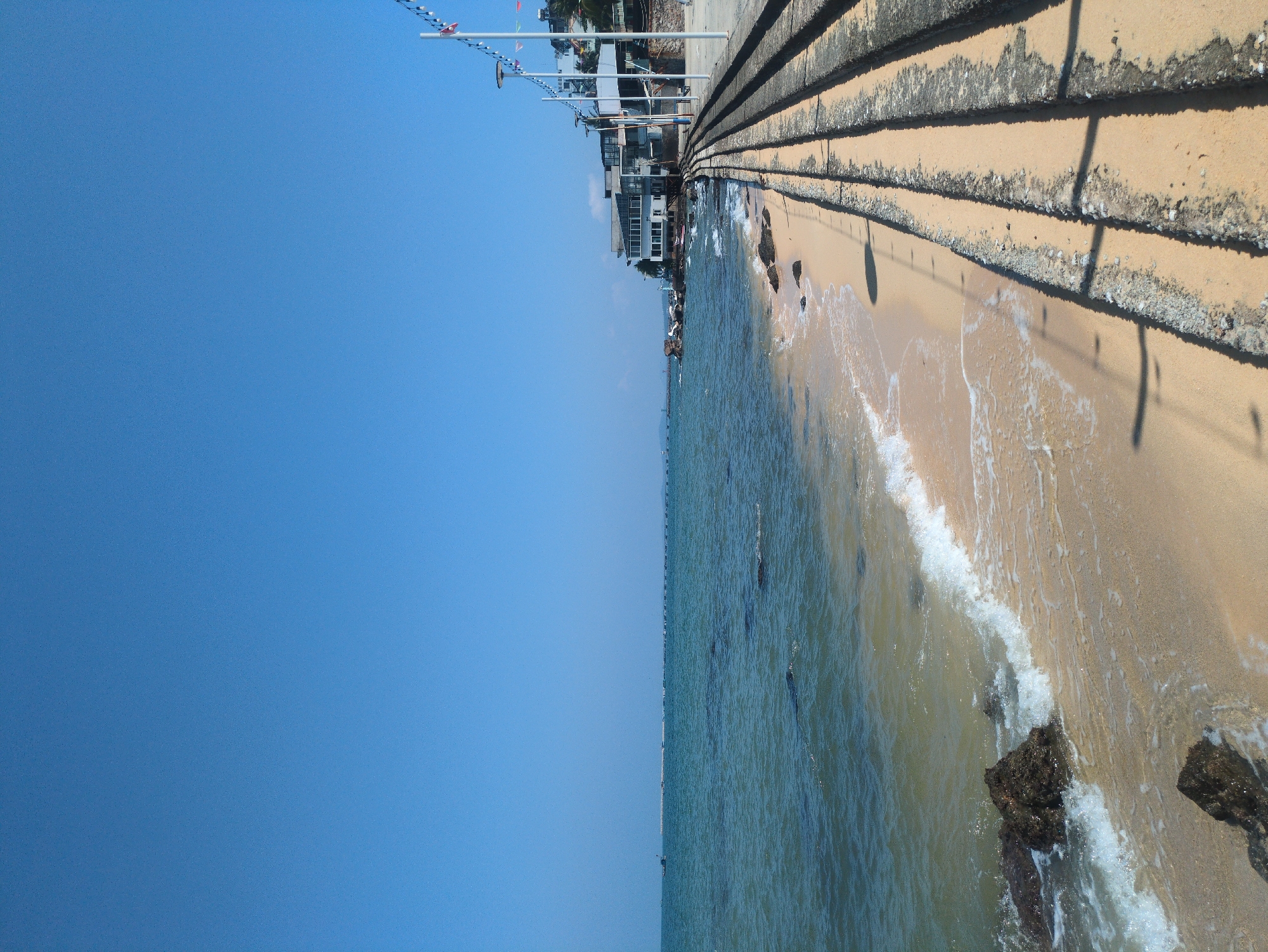 The image shows a beach scene in Phú Quốc, Vietnam, photographed on a bright, sunny day. The perspective is a horizontal orientation, with a split view of the sky and the coast. The clear, light blue sky occupies the left half of the image. On the right, the coastline consists of a sandy beach where gentle waves wash ashore. The beach is backed by a concrete sea wall with steps leading up to a promenade or street lined with buildings. The buildings nearest to the sea wall appear to be multi-storied and likely commercial or residential. Light posts extend over the beach area from the buildings. The water is a mix of light green and turquoise colors, reflecting the sunlight. Rocks are scattered along the shoreline. The scene is still and suggests a tranquil setting. No people are visible in the frame.