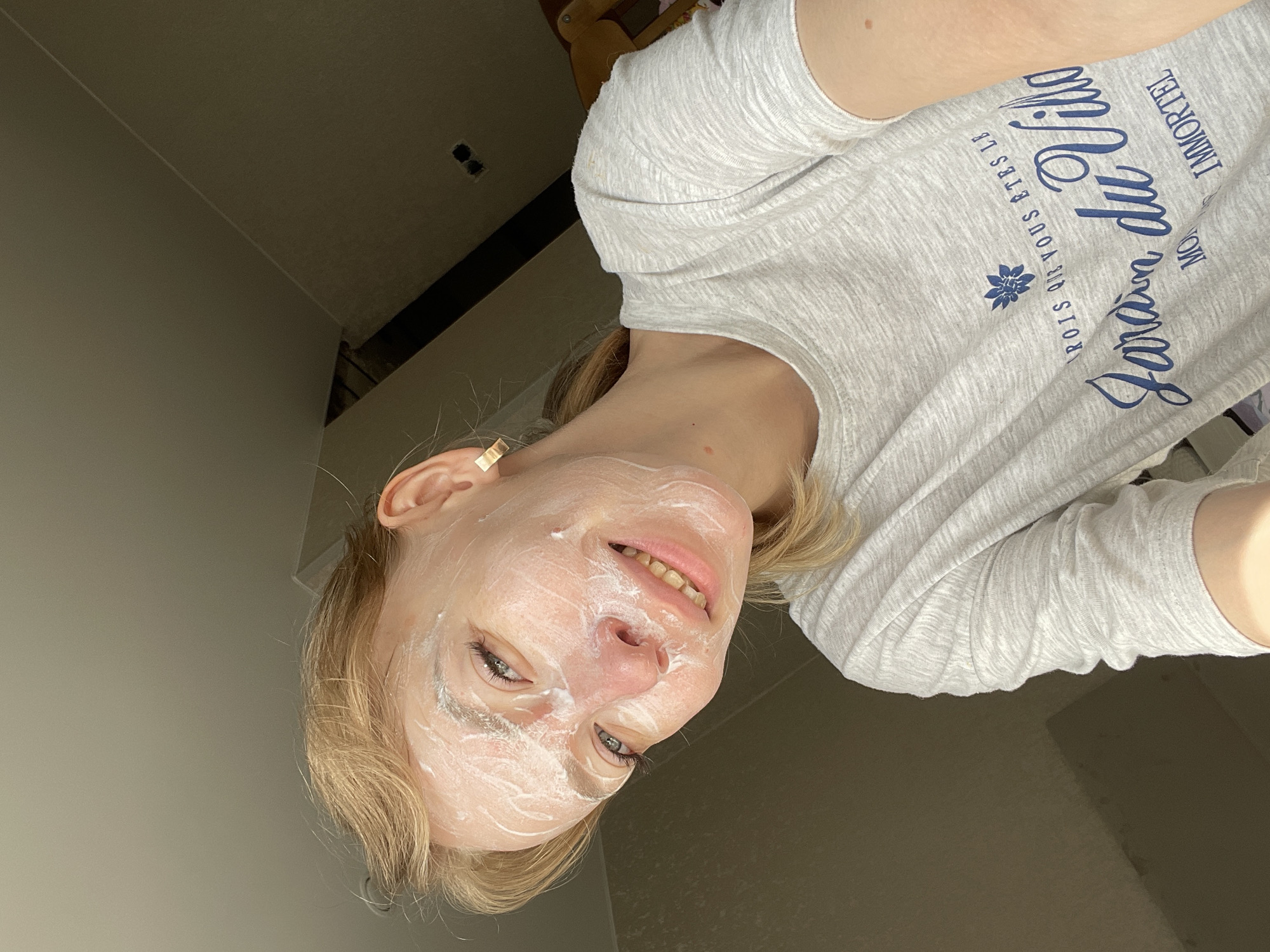 The image shows a woman with blonde hair, lying down and facing the camera. She is wearing a light grey t-shirt with dark blue lettering. There is a white facial mask applied to her face. The background includes a wall and part of a room. There is no information available to determine the location in Perm, Russia.