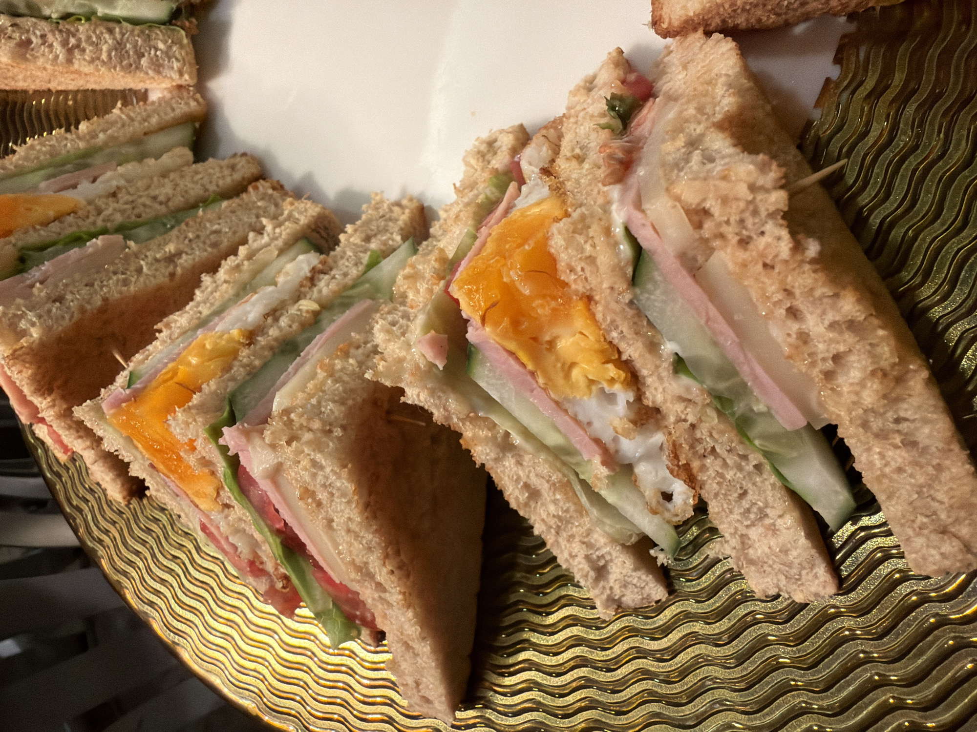 This image shows a close-up, low-angle shot of several club sandwiches cut into triangles and arranged on a wavy, golden platter. The sandwiches appear to be made with whole wheat bread and contain layers of ham, a fried egg with a visible yolk, lettuce, tomato, and cucumber. The platter is placed on a white surface. The background is blurred but suggests an indoor setting with vertical elements. The image is well-lit, highlighting the textures and colors of the food. The precise location in Thuận Hóa Ward, Vietnam is not discernible from the visual information provided.