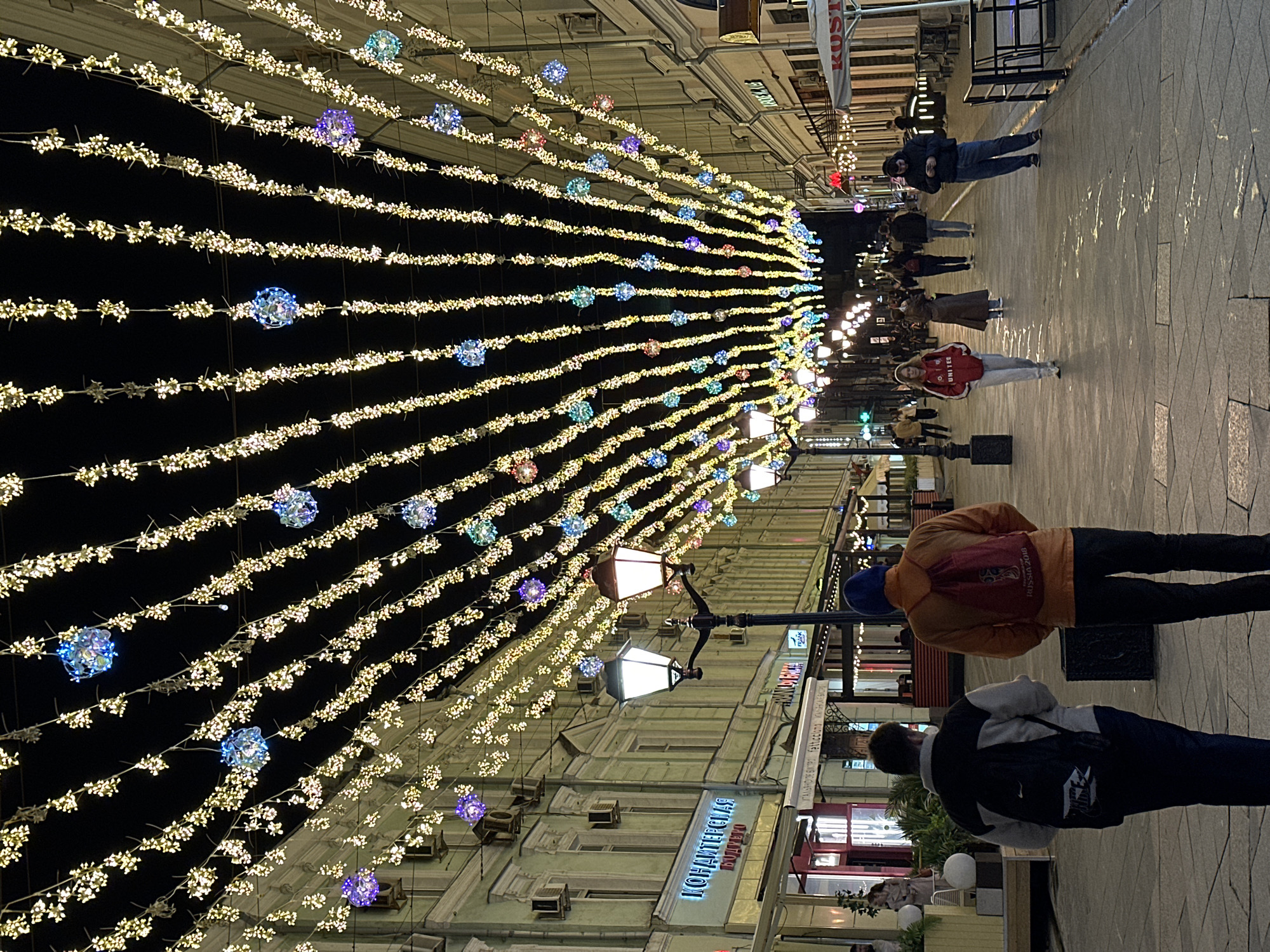 The image depicts a pedestrian street in Moscow, Russia, illuminated with decorative lights strung overhead. These lights form a dense canopy, with rows of small white lights interspersed with larger, colorful spherical lights. A street lamp is visible on the right side of the frame.
People are walking along the street. Some individuals are clearly visible, including a person wearing an orange jacket with a red backpack and another wearing a black jacket with a white logo, possibly Nike. Others are more distant and appear as blurred figures.
Buildings line the street on either side. The signage is visible on some buildings, including a sign with Cyrillic script: "КОНАПИТЕССКАЯ ВОЛТЕЛЯ." A "KOSTI" sign is also visible further up the street.
The street is paved with large stone blocks. The overall impression is of a lively urban street scene, likely during the evening or night.