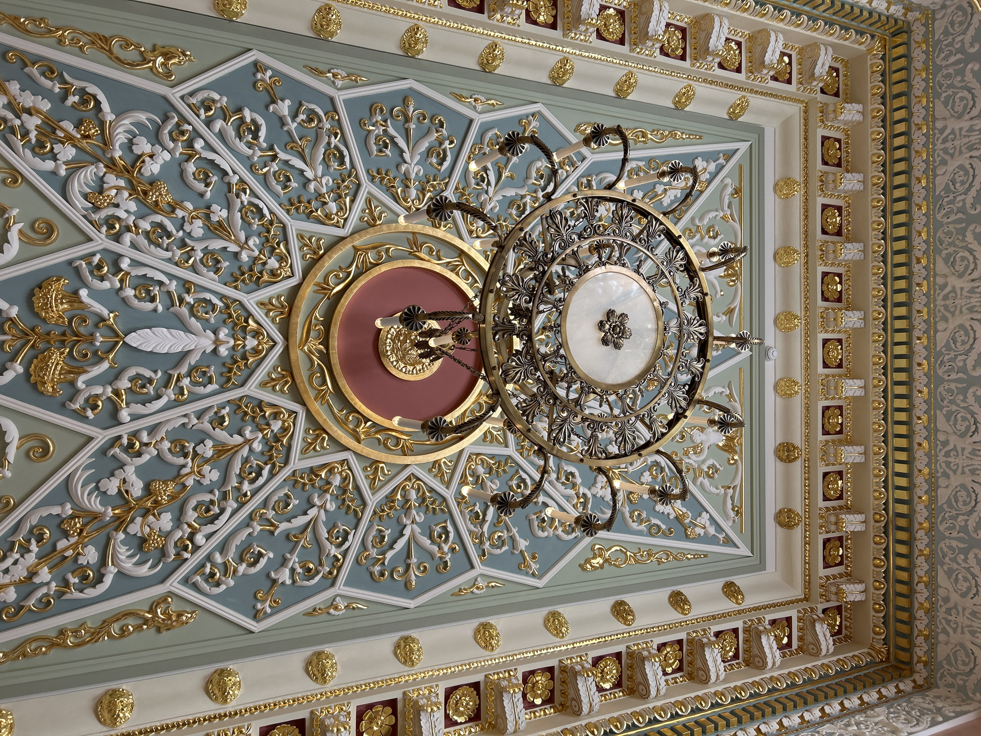The image shows a highly ornate, decorated ceiling, likely in a grand interior space. The ceiling features intricate plasterwork with gold accents, painted in shades of light blue, cream, and gold. Geometric patterns and floral motifs are prominent. A large, dark metal chandelier with multiple candle-like lights hangs from the center of the ceiling. The chandelier has a circular frame with decorative metalwork and a central frosted glass element. The overall impression is one of opulence and classical architectural style. The image does not contain any people or visible actions. There is no information within the image itself to indicate its location in Добасненскі сельскі Савет, Belarus.
