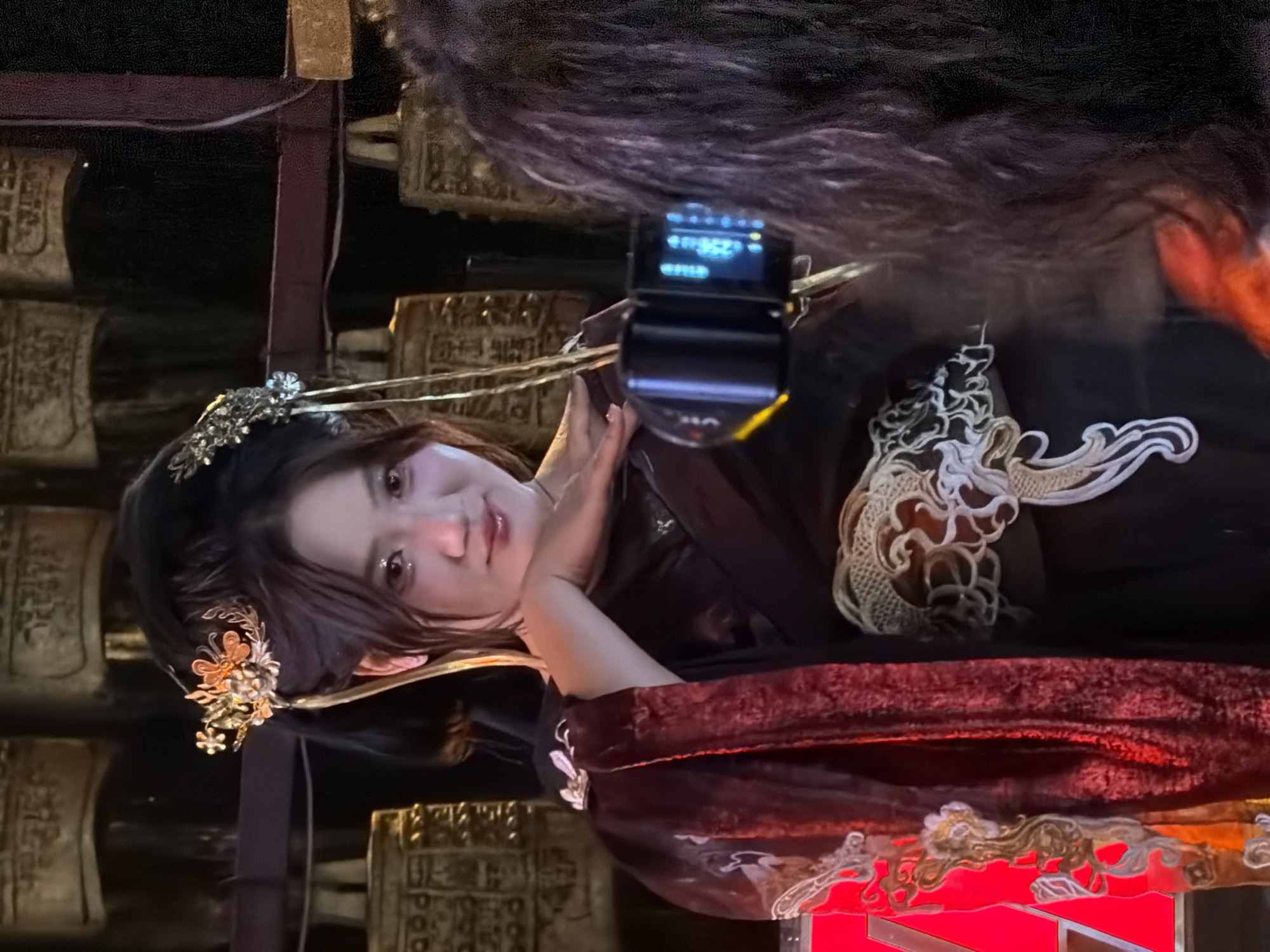 The image shows a young woman in traditional Chinese attire, posing in front of a display of bells. She has her hair styled up with ornate hair accessories and is wearing a dark garment with red and gold accents. Her left hand is resting on her chin, and she is looking directly at the camera. In the foreground, the back of another person's head and shoulder are visible, holding a camera to take a photo of the woman. The background consists of several large, ornate bells, likely part of a cultural exhibit or performance. The overall setting appears to be indoors, possibly a museum or performance hall. The lighting is dim, with a spotlight on the woman. Based on the presence of the bells and the woman's attire, the location is likely related to ancient Chinese culture. The Yuzhong District in China is known for its historical and cultural sites, making it a plausible location for such a scene.