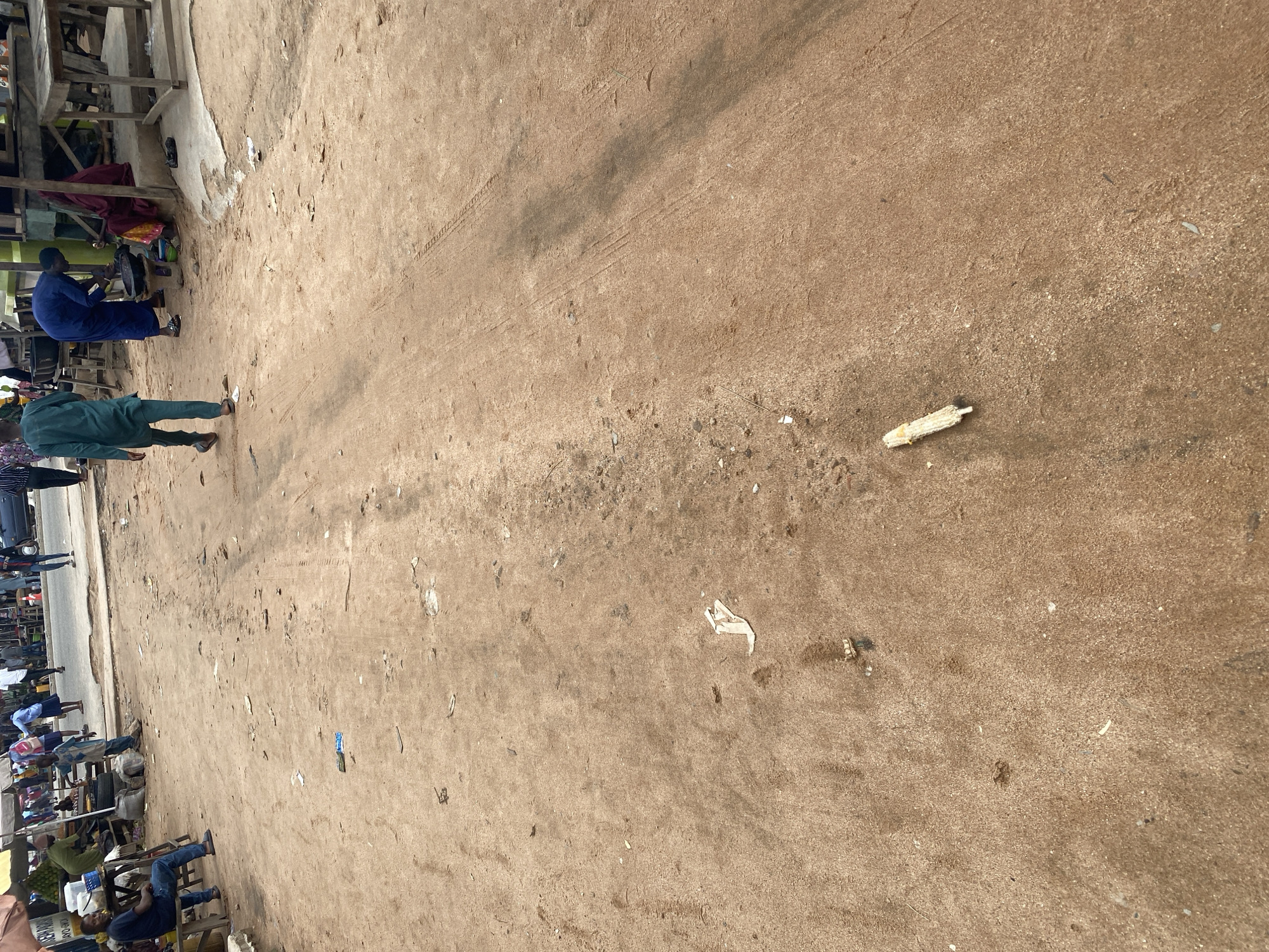 The image depicts an outdoor scene in Jalingo, Nigeria, likely a marketplace given the presence of stalls and a road. The ground is covered in sandy dirt, with some scattered debris and tire tracks visible, suggesting it's a well-trafficked area.
In the foreground, a person in a green outfit stands facing away from the camera, with another person in a blue robe seated at a stall to their left. Several other individuals are visible further down the road, some seated at stalls and others standing. The stalls appear to be makeshift structures, with wooden frames and possibly tarpaulin coverings. There are goods displayed at some of the stalls, including what looks like produce.
The lighting suggests it is daytime, possibly late morning or afternoon. The weather appears to be dry. The overall atmosphere seems relaxed, with people engaged in their daily activities. There are no clear indicators of specific emotions, but the scene suggests a typical bustling market environment. No discernible text is visible in the image. A piece of what looks like corn cob is present on the ground in the mid-right section of the image.