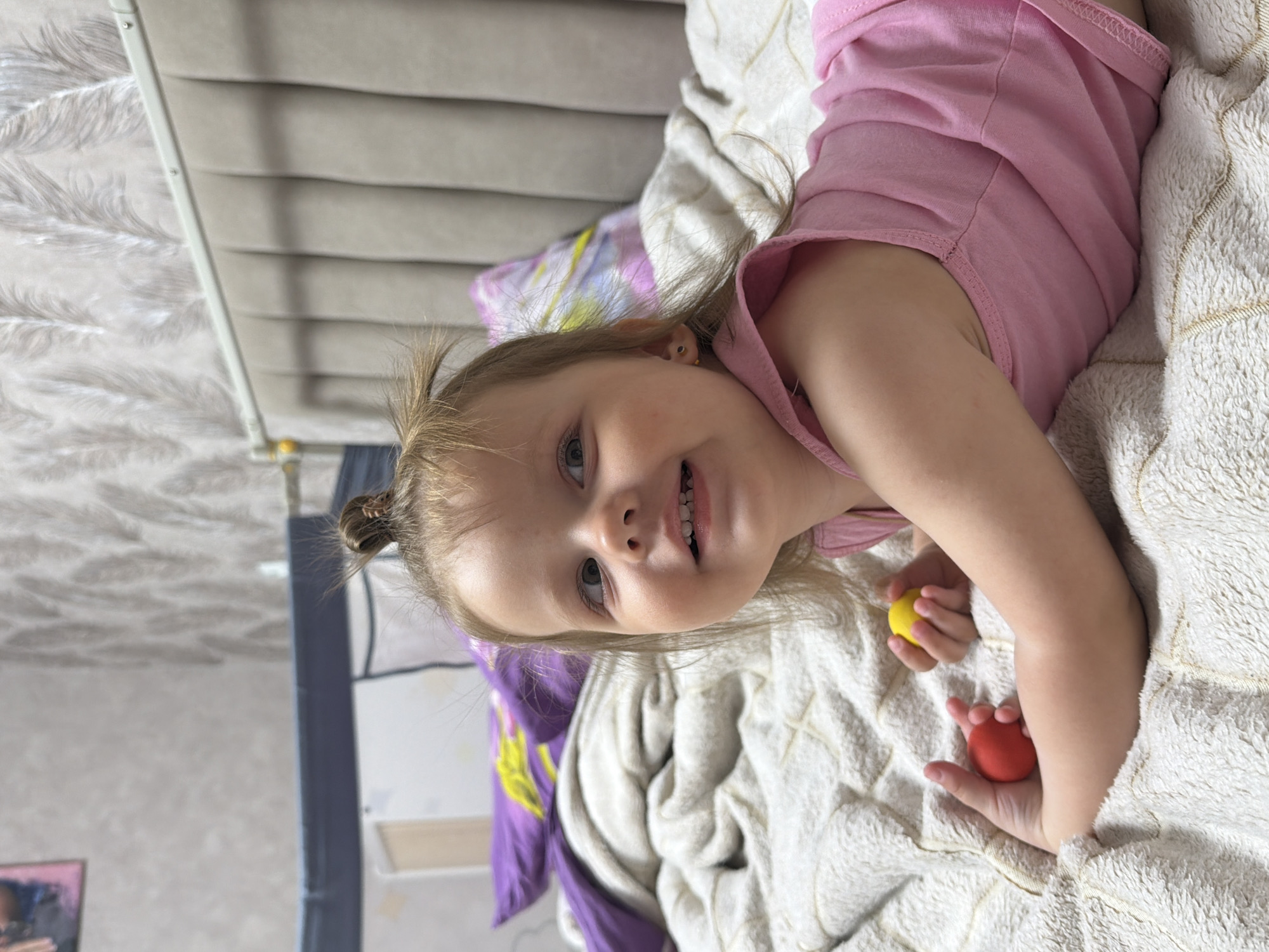 A young child lies on a bed. The child is wearing a pink top. In the child's hands are a red ball and a yellow ball. The bed has a textured white blanket, and a purple and patterned pillow is also visible. The headboard behind the bed is gray. The location appears to be indoors, with a wall visible in the background. The location is not confirmed to be in the city of Reshetnikovo, Russia.