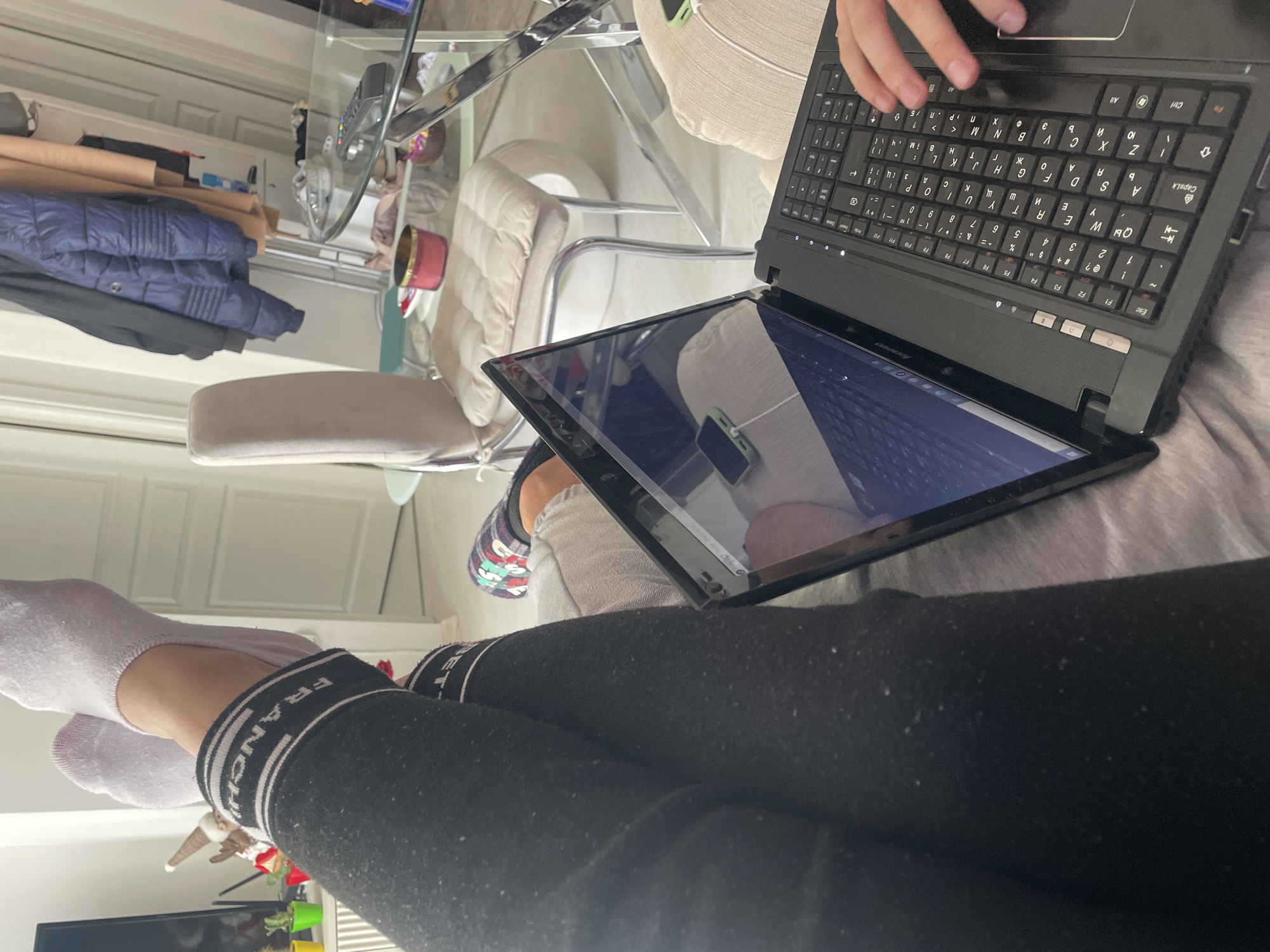 The image shows a person seated, using a black Lenovo laptop. The laptop is open, and the person's hand is visible on the keyboard. They are wearing black leggings with the word "FRANCH" repeated on the waistband and white socks. The background includes a glass table, a white cushioned chair, and a glimpse of a room interior with a white door and some clothing items. The scene is likely indoors. The specific location in Sofia, Bulgaria, cannot be determined from the image alone.