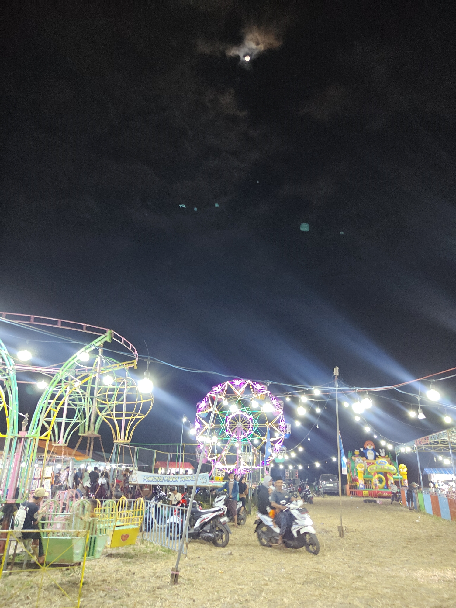 This photograph depicts a nighttime scene at a fairground, likely in Suling Wetan, Indonesia. The sky is dark, with the moon partially obscured by clouds, and illuminated by bright, directional beams of light emanating from unseen sources, possibly stage lights or spotlights. In the foreground, dry grass covers the ground.
Several amusement rides are visible. To the left, a large, colorful structure with intricate metalwork and hanging lights appears to be a swing ride or a similar attraction. Further into the background, a Ferris wheel adorned with multicolored lights dominates the skyline. To the right, a large inflatable bounce house featuring cartoon characters is set up.
Numerous people are present, many standing around the rides or walking through the fairground. Several motorcycles are parked or being ridden by individuals. In the mid-ground, a group of people are gathered near a concession stand or game booth. A banner with text, partially obscured, is also visible. The overall atmosphere suggests a lively and festive event.
