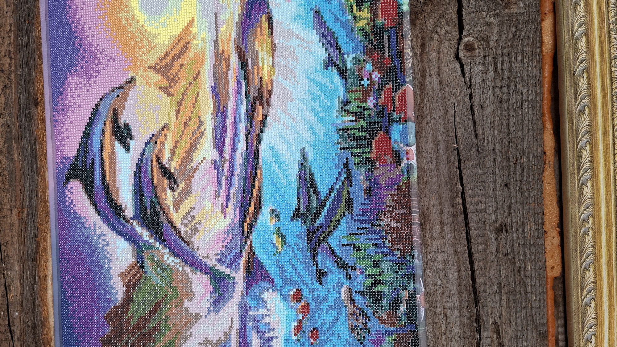A diamond-painted canvas featuring two dolphins leaping above a stylized landscape of trees and water, partially framed, rests against a weathered wooden surface. The painting is composed of numerous small, colorful diamond-shaped beads. The frame is ornate and gold-colored. The wooden surface is rough and dark with a visible knot.
There is no information that allows a determination of location or city of Большой Букор, Russia.