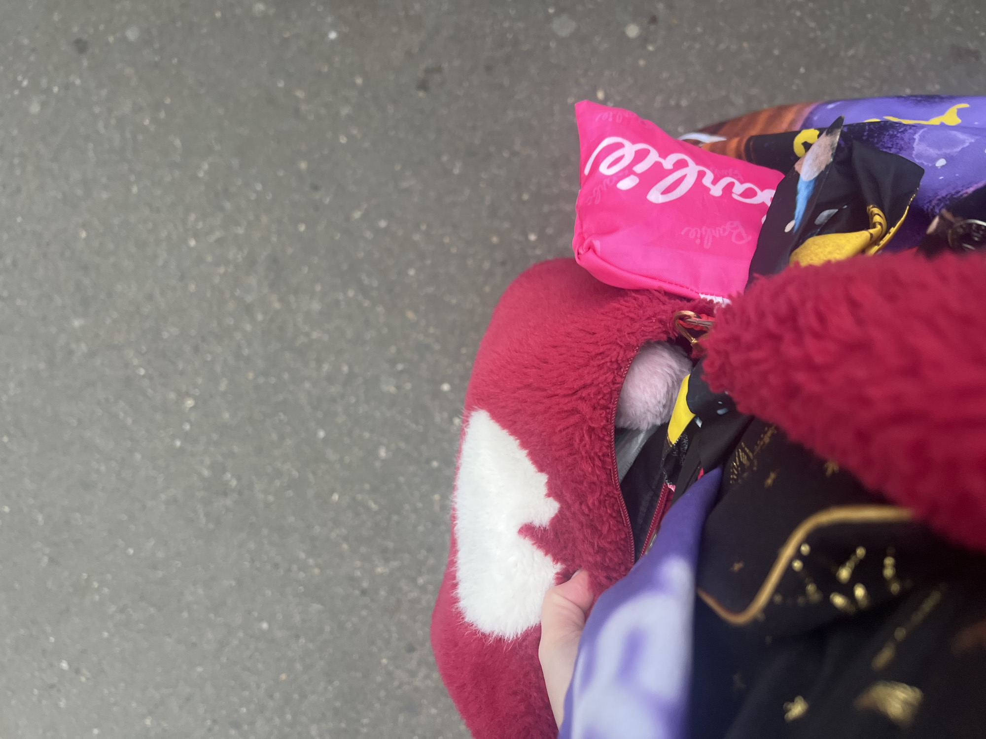 The image presents a close-up, high-angle view of a collection of colorful items, likely bags or soft goods, set against a blurred, textured grey background, indicative of an outdoor concrete or asphalt surface. The scene is illuminated by bright, even light, suggesting daytime in Tbilisi, Georgia.
Prominently featured is a vibrant pink pouch or small bag with "Barbie" written in large white script across its surface, with smaller "Barbie" text visible beneath. Adjacent to this is a fluffy, deep red item, possibly a bag or garment, distinguished by a contrasting white shape on its side, which could be a heart or an abstract design. A hand with light skin is visible on the right, appearing to hold or interact with the red fluffy item.
Among these, other textiles can be seen: a segment of purple fabric with yellow patterns, and a darker material, possibly black, adorned with glittering gold specks and wavy lines. A small, soft, light pink or white fluffy object peeks out from an opening, suggesting it is tucked inside one of the bags. The composition implies a casual moment, perhaps of someone carrying personal belongings while outdoors.
