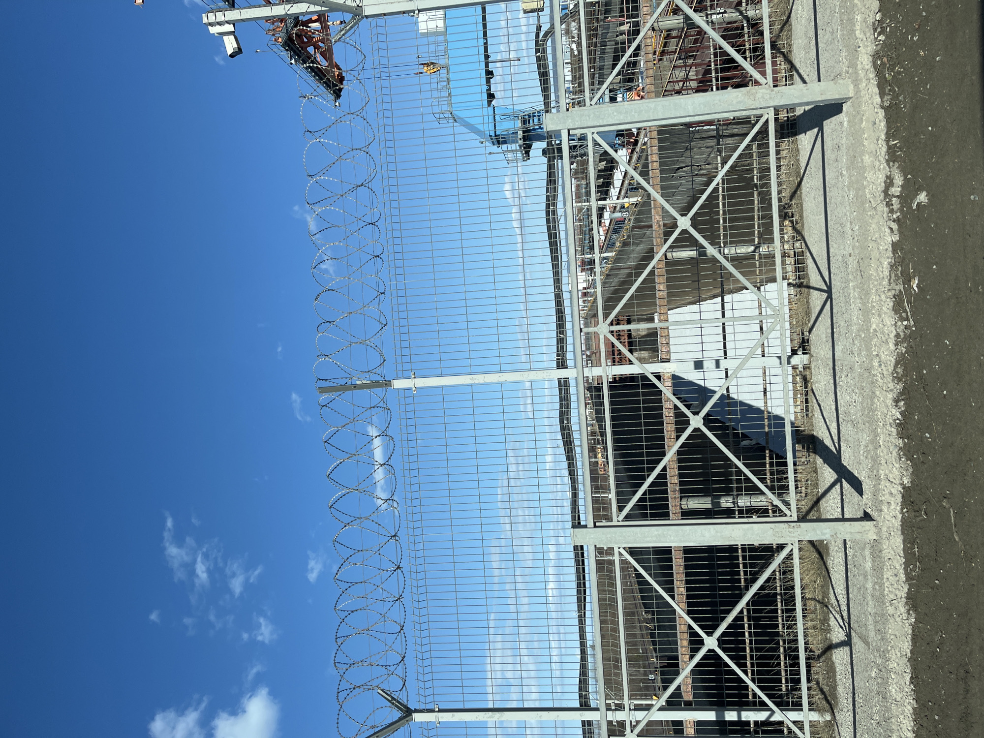 The image depicts a security fence with barbed wire in Chaikovsky, Russia, against a clear blue sky with some clouds. The fence is made of metal mesh and a frame reinforced with diagonal supports. It runs parallel to a concrete curb. Behind the fence, glimpses of industrial infrastructure are visible. These include metal beams, pipes, and structures partially obscured by the fence. An antenna is positioned in the upper-left of the frame.