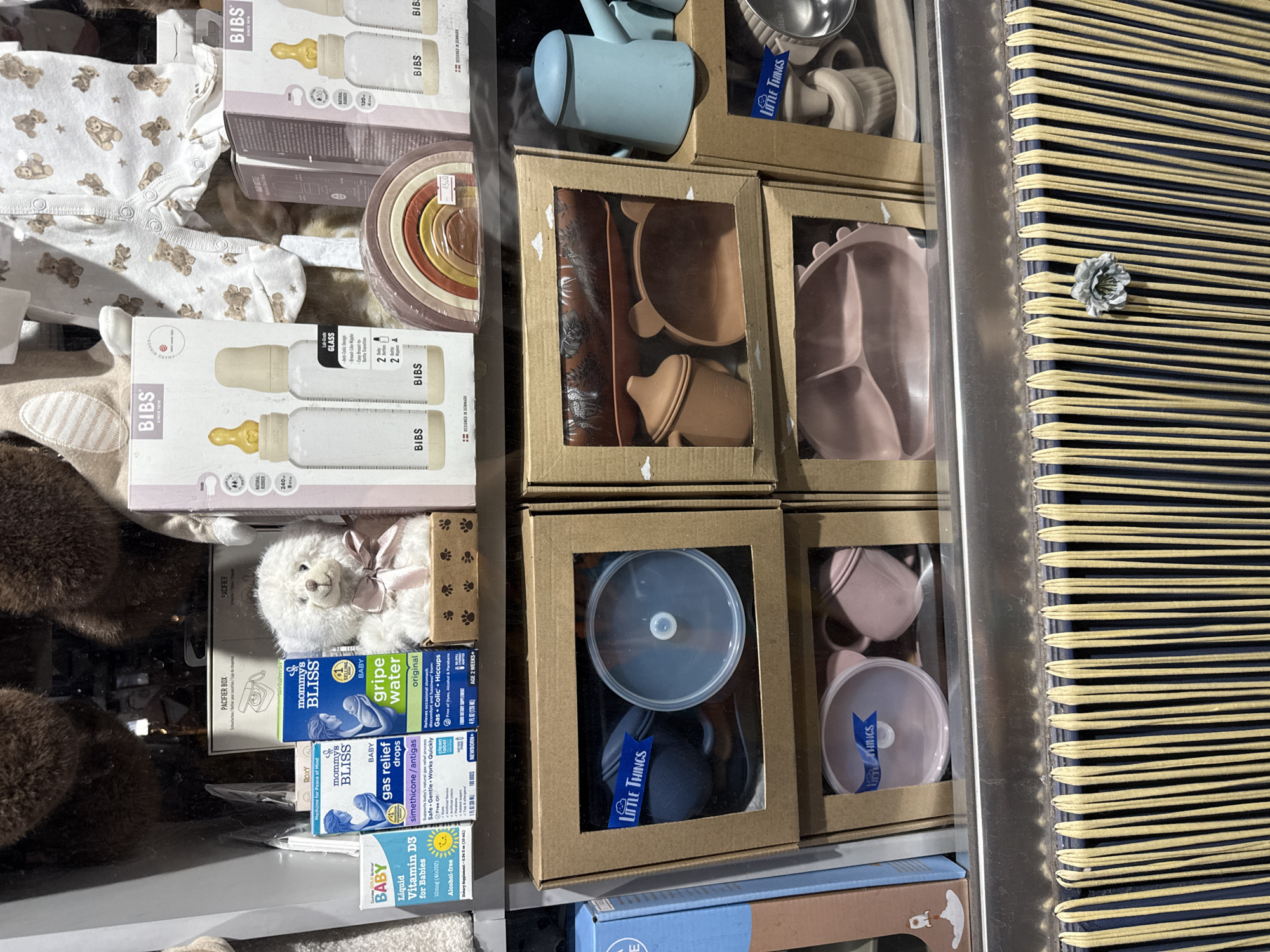 This image depicts a display of baby products in a shop window. The products include baby bottles, a teddy bear, pacifiers, baby formula, gripe water, and silicone tableware. The display is neatly arranged with cardboard boxes and clear plastic packaging. The overall impression is one of a well-stocked baby store. The location is not discernible from the image.