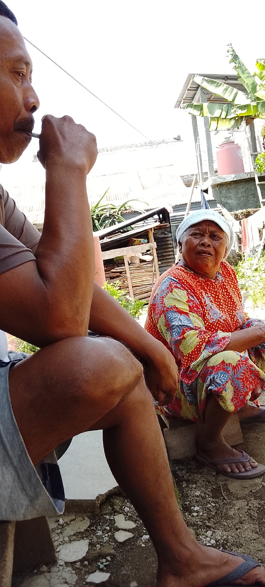 In Padang Dalom, Indonesia, an older woman with a light-colored head covering and an orange shirt with a floral pattern sits on a wooden plank. She appears to be looking towards the left. Beside her, a man is visible from the chest down, with his left arm resting on his knee. He is smoking and looking to the left. The background shows a rustic environment with tin roofs, vegetation, and a water tank.