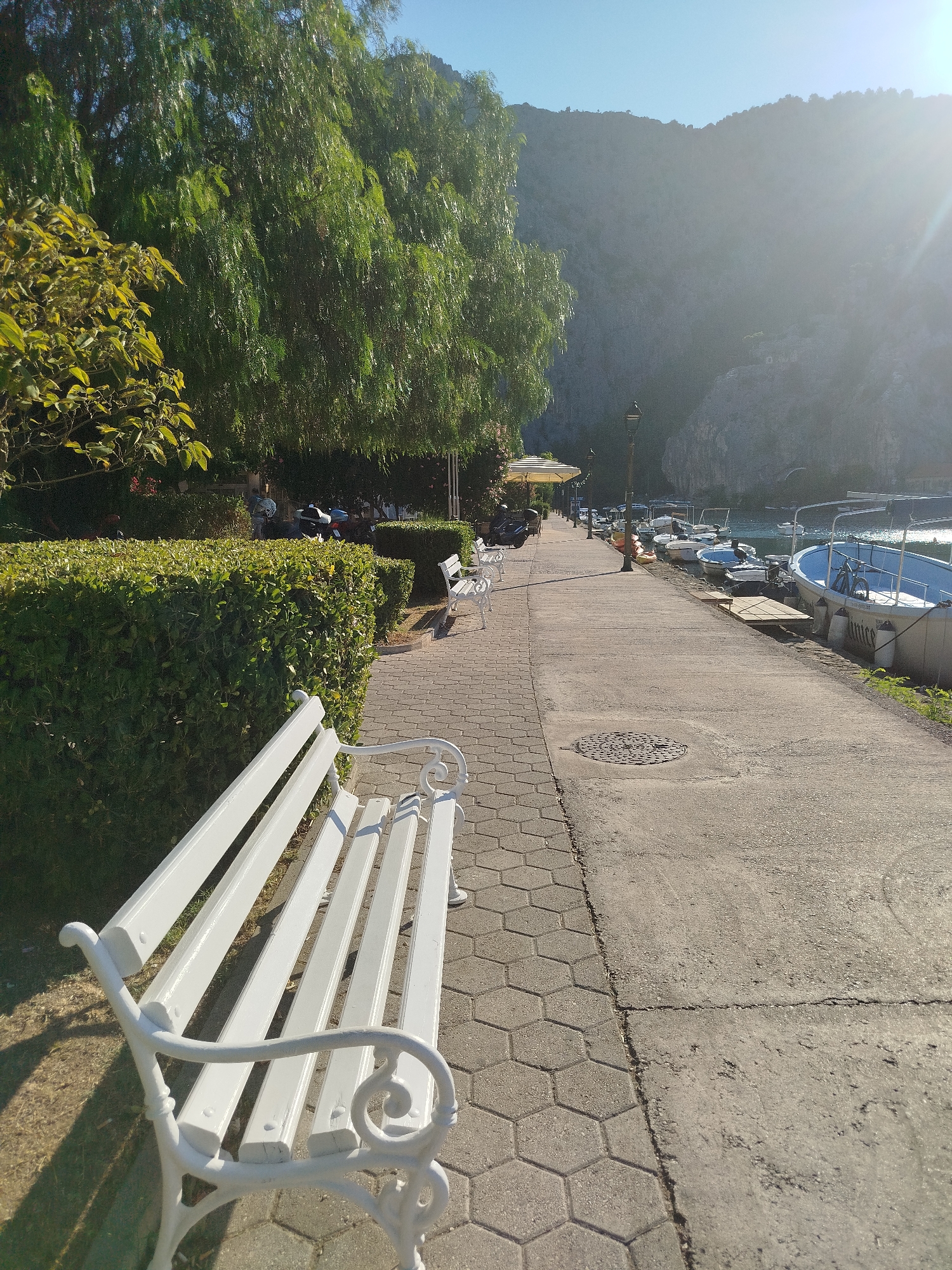 The image depicts a sunlit waterfront promenade in an unknown city. In the foreground, a white ornate bench is positioned on a hexagonal paved path. To the left of the path, a well-maintained green hedge lines the walkway, behind which are trees with drooping branches and some yellow-leafed foliage. Beyond the hedge, several white benches and a parked motorcycle are visible.
The promenade itself is made of concrete and stretches into the distance, lined with lampposts. On the right side of the promenade, a waterway is visible, with numerous boats docked along the edge. The boats are of various sizes and colors, including a prominent blue and white boat in the right foreground. In the background, a large, rugged mountain range rises, bathed in the bright sunlight, with some trees and structures appearing on its slopes. The sky is clear and blue, with a lens flare from the sun casting a bright glow over the scene.