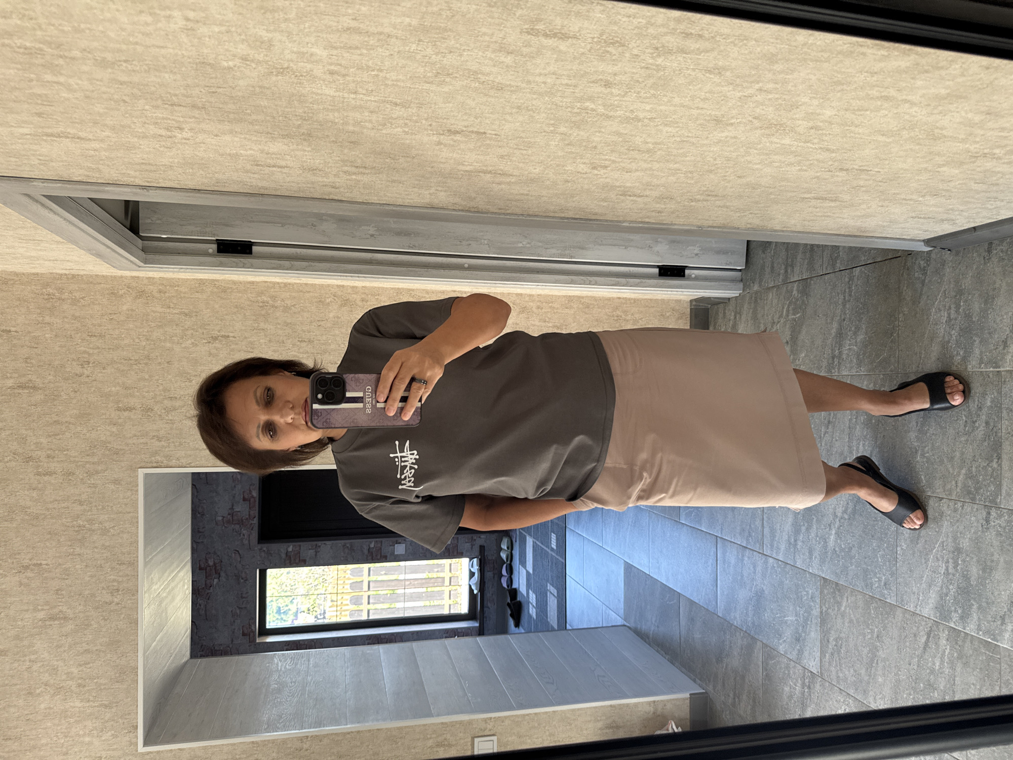 A middle-aged woman with brown hair is standing in a hallway and taking a selfie in a large mirror. She is wearing a dark grey t-shirt with a white graphic print and a light brown skirt. She has black sandals on and is holding a black phone with a purple case in her right hand. The hallway has grey tiled floors and textured beige walls. To the left of the woman is a doorway leading to another room, and to her right, a closed door with a light grey frame. In the background, through an open doorway on the left, a section of brick wall and a window with a fence are visible. Several pairs of slippers are neatly arranged on the floor in the background.