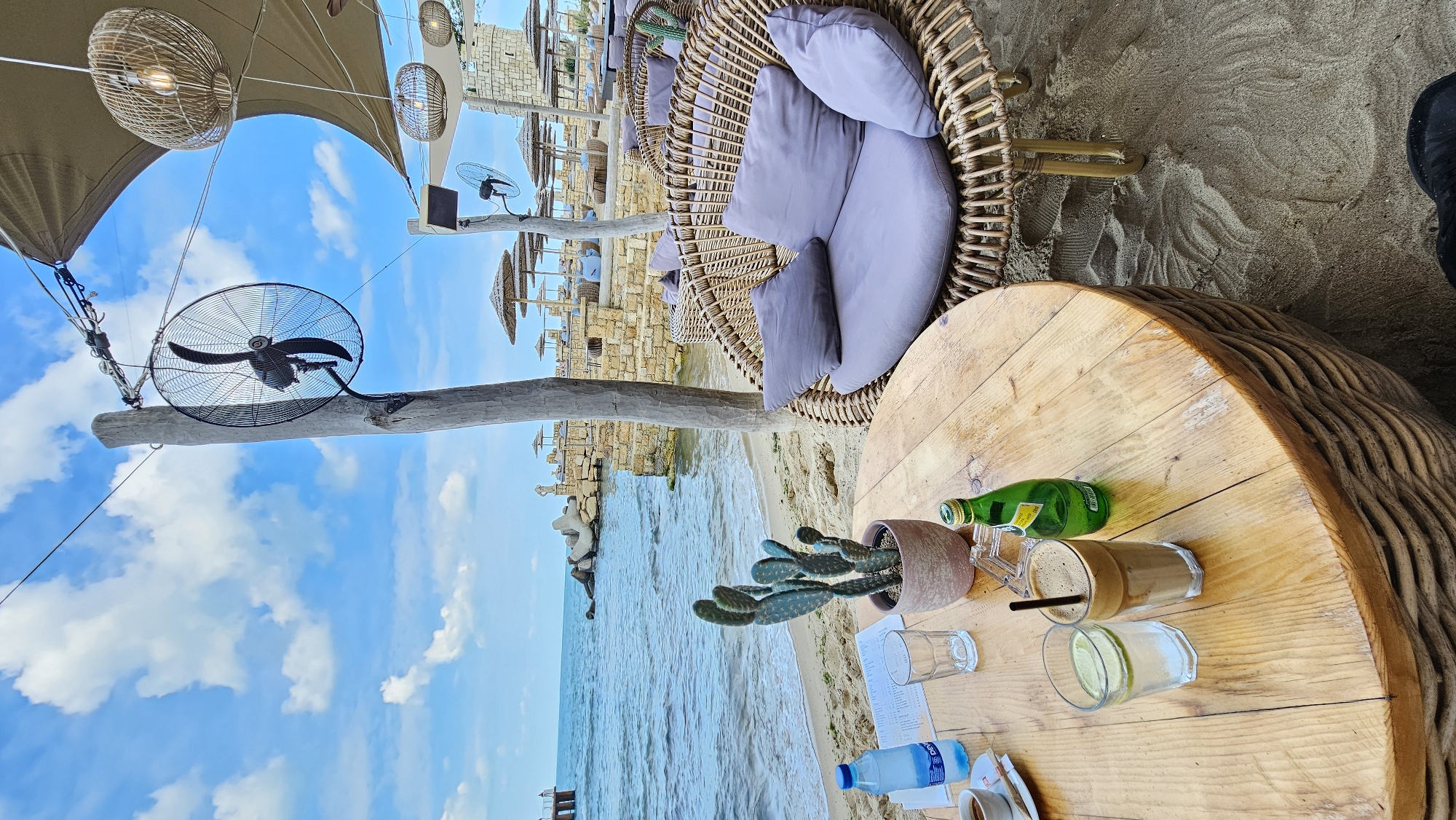 The image captures a relaxed outdoor scene, likely in Varna, Bulgaria, as suggested by the coastal environment and architecture. The perspective is from a seated position at a round wooden table. On the table are beverages (iced coffee, water, sparkling water), a potted cactus, and disposable tableware. A woven wicker chair with plush cushions is adjacent to the table, suggesting a comfortable seating area. The beach with light sand stretches out towards the blue water, and stone structures (likely a protective wall or part of a seaside establishment) line the coast. Beach umbrellas and sunbeds are visible in the background. Above, there is a large overhead fan and a sunshade, indicating a shaded area for patrons. The sky is blue with some clouds, suggesting a sunny day. No people are clearly visible in the foreground, although the presence of the table setting implies human activity.