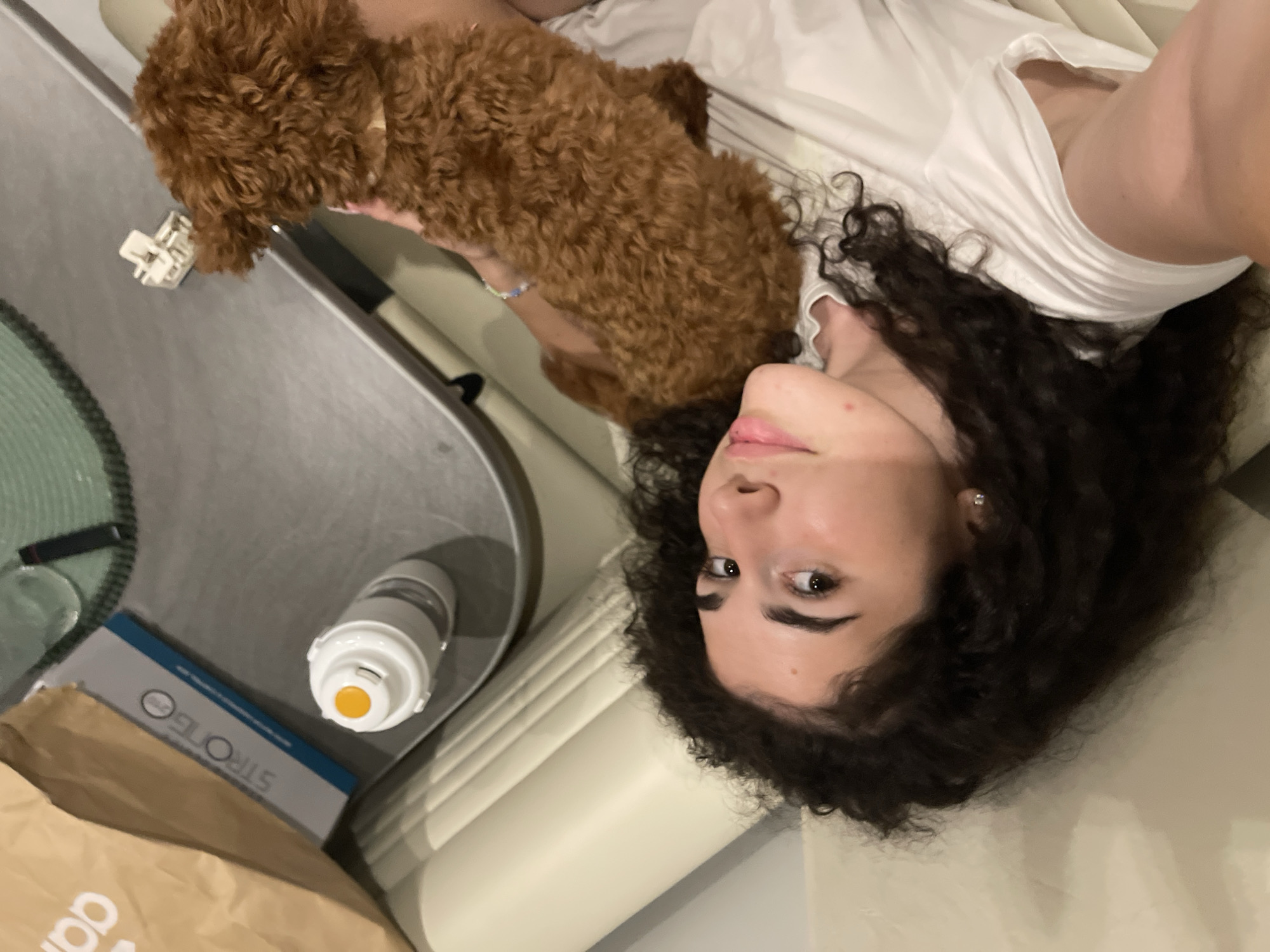 This image, captured in Koprinka, Bulgaria, presents an indoor scene featuring a young woman and a small dog in a relaxed setting.
A young woman with dark, curly hair and light complexion is depicted lying on a light-colored upholstered surface, possibly a couch or bed, positioned towards the left side of the frame. Her head is angled to the left, and her face is looking towards the right side of the image. She has dark eyes, distinct eyebrows, and her lips are slightly parted. A small, silver stud earring is visible in her left earlobe. She is wearing a white short-sleeved t-shirt. Her right arm, adorned with a blue and white beaded bracelet, extends towards the right, supporting a small, reddish-brown dog.
The dog, appearing to be a poodle or poodle mix due to its dense, curly fur, is nestled on the woman's chest or shoulder area. It wears a light-colored collar and is facing away from the camera, looking towards the upper right of the frame.
In the background, positioned on a dark gray or metallic table surface behind the woman's head, several objects are visible. These include a white water bottle with a clear lower section and a prominent orange push-button mechanism on its cap. Next to the bottle is a rectangular item, possibly a book or document, displaying "STRONGO" in white text on a blue band. Partially obscured by this item is a brown paper bag with white text, resembling part of an "adidas" logo ("ad" is visible). Further to the upper right on the table, near the dog's head, a small, light-colored claw hair clip is present. A textured green circular or oval object, possibly a mat or coaster, is partially visible on the far top left of the table.
The lighting is soft and even, suggesting an indoor environment.