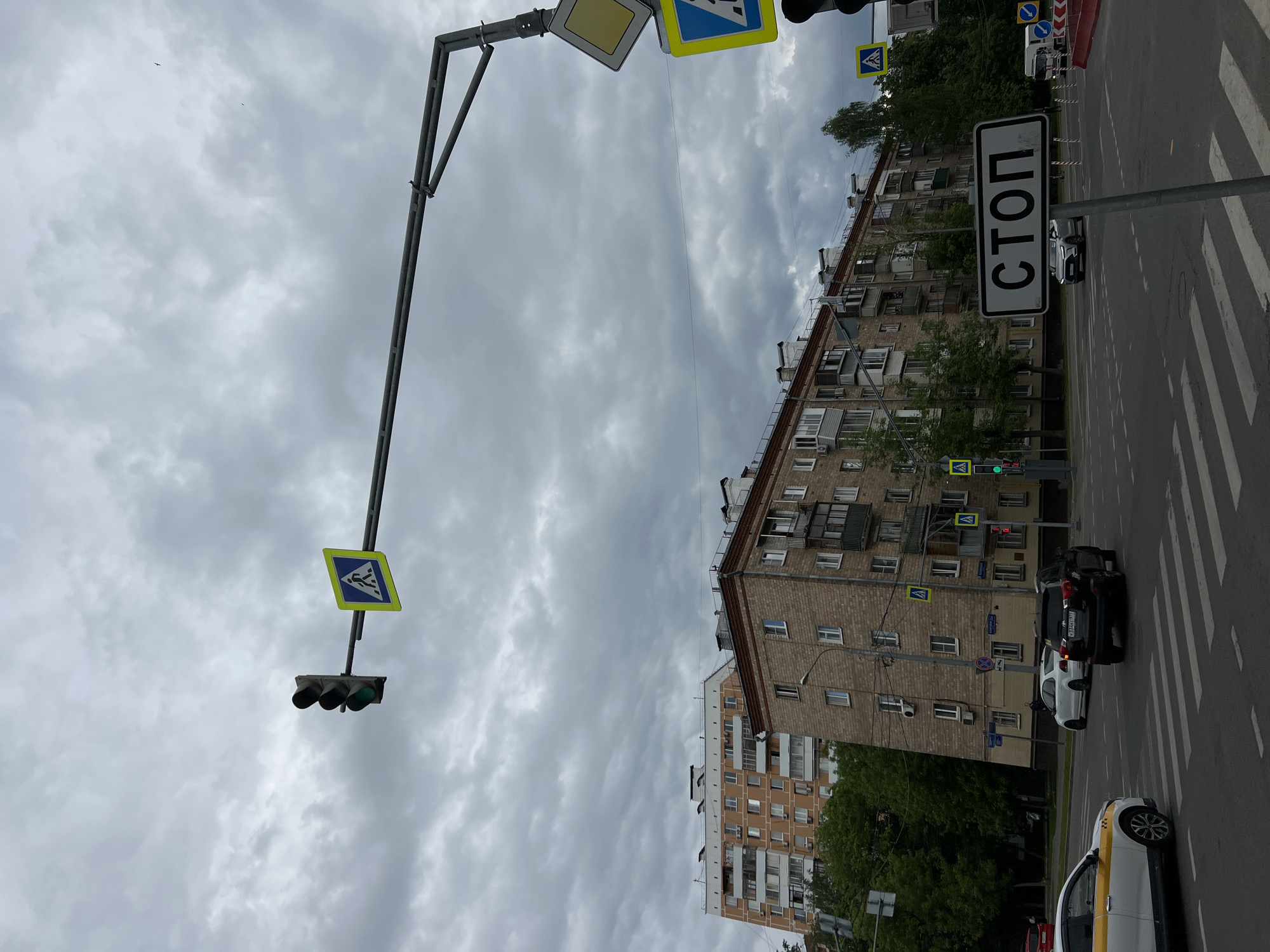 The image shows a street scene in Moscow, Russia. The street is lined with buildings, and there are several cars parked on the side of the road. There is a pedestrian crossing with a crosswalk sign, and a traffic light showing a green signal. A "STOP" sign is visible. The sky is cloudy and overcast.