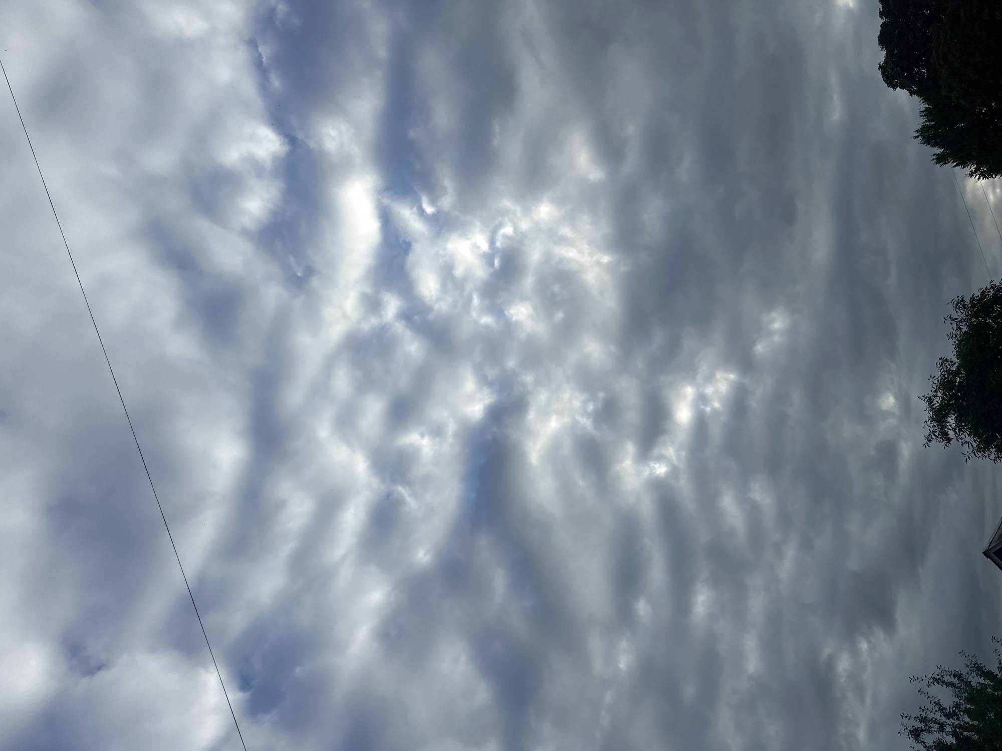 The image shows a view of the sky with dark, textured clouds that obscure much of the sun. A thin, dark wire runs diagonally across the upper portion of the frame. In the lower part of the image, the tops of dark green trees are visible, along with the peak of a dark, angular roof, suggesting a residential area. The overall impression is of an overcast or stormy sky. This scene is not specific to Khmilnyk, Ukraine, and contains no identifiable landmarks or specific features that would indicate that location.