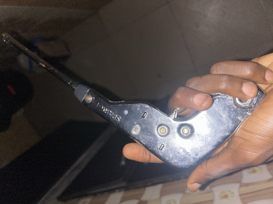 A hand holds a spark lighter against a blurred background. The spark lighter, labeled "SPARK-I", is silver-colored with a long, thin, black nozzle. Two buttons are visible, likely used to generate the spark. The hand has dark skin and is gripping the lighter firmly. The background is out of focus, but suggests an indoor setting with patterned surfaces. The lighting is dim, making details less clear.
