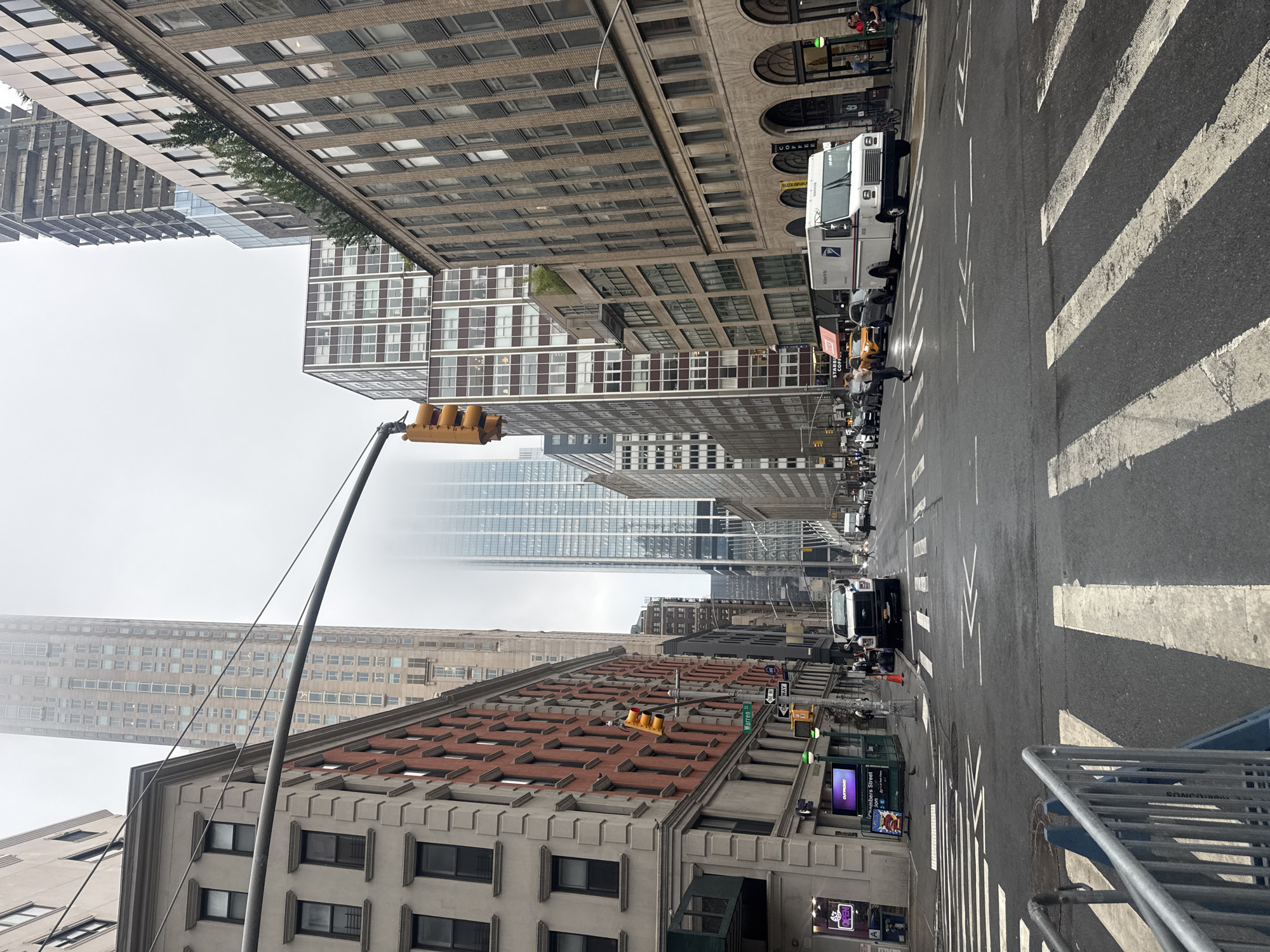 This is a street-level view of a city intersection in New York, United States of America. The scene is dominated by tall buildings on all sides, with a gray, overcast sky. The street is wet, suggesting recent rain. There are multiple crosswalks with white stripes on the asphalt.
In the foreground, there is a metal barricade on the left, partially obscuring the view of the sidewalk. Several cars are visible in the distance, including a white postal service truck in the middle of the intersection, stopped at a red traffic light. A few pedestrians are present, walking on the sidewalks and crossing the street.
Several traffic lights are visible, with one showing yellow. Street signs indicate "Warren St" and "One Way". Buildings vary in architectural style, with some featuring brick facades and others modern glass exteriors. The overall atmosphere is urban and somewhat muted due to the weather.