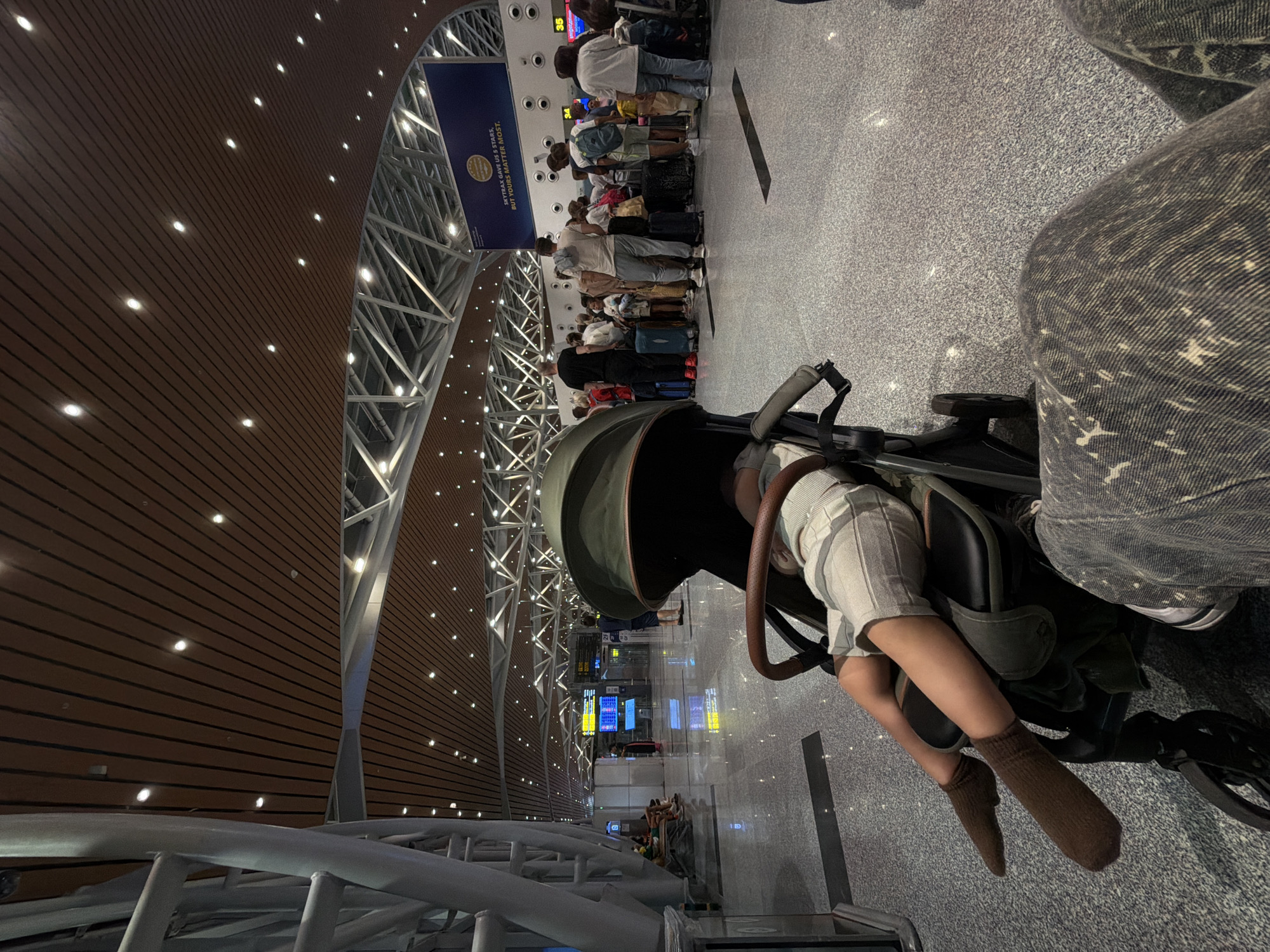 This image captures a moment within a modern airport terminal, likely in Da Nang, Vietnam. In the immediate foreground, a child or baby is seen resting or sleeping in a dark-colored stroller. The child wears light-colored shorts and brown socks. Adjacent to the stroller, a leg clad in distressed denim jeans is partially visible, indicating the presence of an adult, possibly a parent, viewing the scene from a low perspective.
Further back, a long queue of people, predominantly adults, stands patiently. They are accompanied by various pieces of luggage, including suitcases and backpacks, lined up on the light grey, speckled floor. Above the queue, digital displays show the numbers "34" and "35," which are likely check-in counter or gate numbers. A large blue sign is prominent in the background, bearing the text: "SKYTRAX GAVE US 5 STARS. BUT YOURS MATTER MOST." and features a golden circular logo, confirming the airport environment.
The terminal's architecture is striking, characterized by high ceilings clad in warm wooden panels embedded with numerous small, bright spotlights. These panels are supported by expansive silver-colored metal truss structures, contributing to a contemporary and spacious aesthetic. The artificial lighting suggests an indoor setting where the time of day is not evident. The overall scene depicts a calm and orderly environment within a busy travel hub.