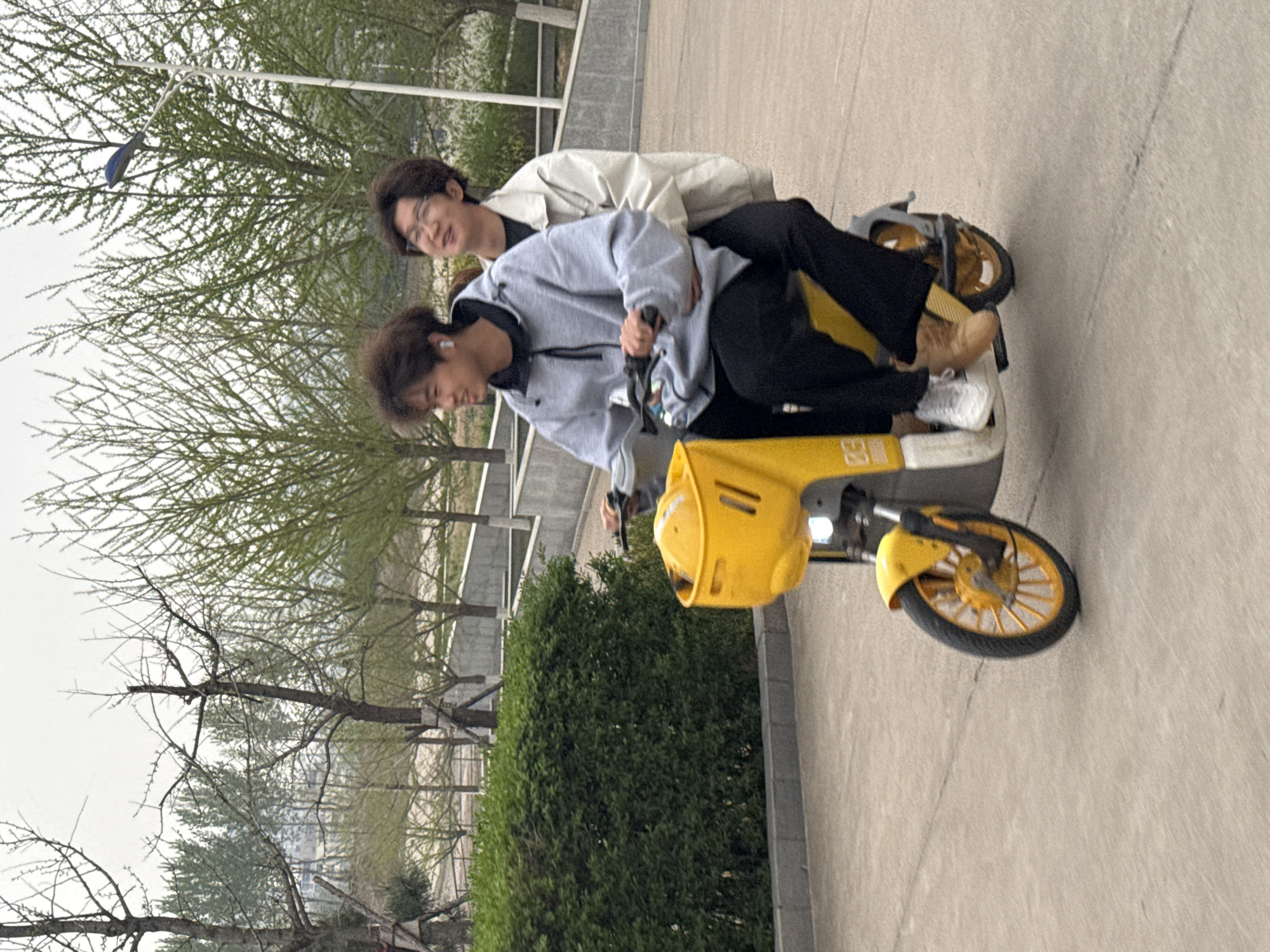 Two young adults are captured riding a yellow electric scooter outdoors. The individual in front, wearing a grey hooded jacket and black pants, is steering the scooter, while the person behind them, in a light-colored jacket and black pants, sits as a passenger. The person in the back wears glasses and smiles towards the camera, while the driver has a subtle smile, looking forward. Both appear to be enjoying the ride. The driver has white earbuds in, and the passenger wears brown shoes.
The setting appears to be a pathway or a road bordered by greenery. Trees with sparse branches and some new green leaves, indicative of early spring, line the background. Lush green bushes are visible to the right. The ground is made of concrete. A street lamp stands tall in the background. The sky is overcast, suggesting daytime with diffuse lighting.
The yellow electric scooter features yellow-spoked wheels and a front basket. Visible text on the side of the scooter reads "E30". Blurry Chinese characters are also present on the front basket, possibly indicating a shared scooter service like HelloBike. The scene conveys a relaxed moment in an urban or suburban green space in Zhongtun, China.