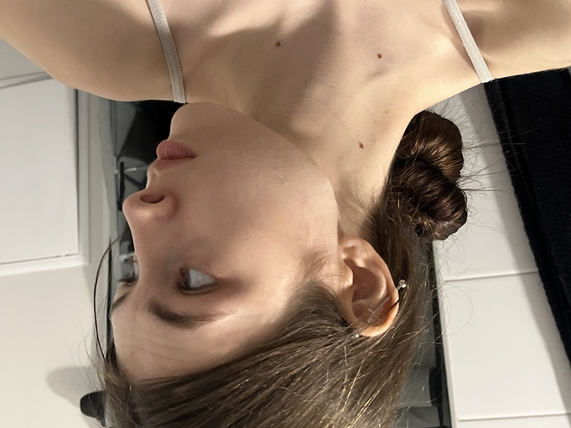 A close-up, angled shot depicts a light-skinned woman's face and upper torso. The woman has brown hair pulled back into a bun, with loose strands framing her face. Her visible features include an eye, nose, lips, and ear, which is adorned with a stud earring. The woman is wearing a light-colored top with thin straps. The background includes a white wall with tile and a dark, textured surface. The image is likely taken indoors. There is no indication of the location being Saint Petersburg, Russia.