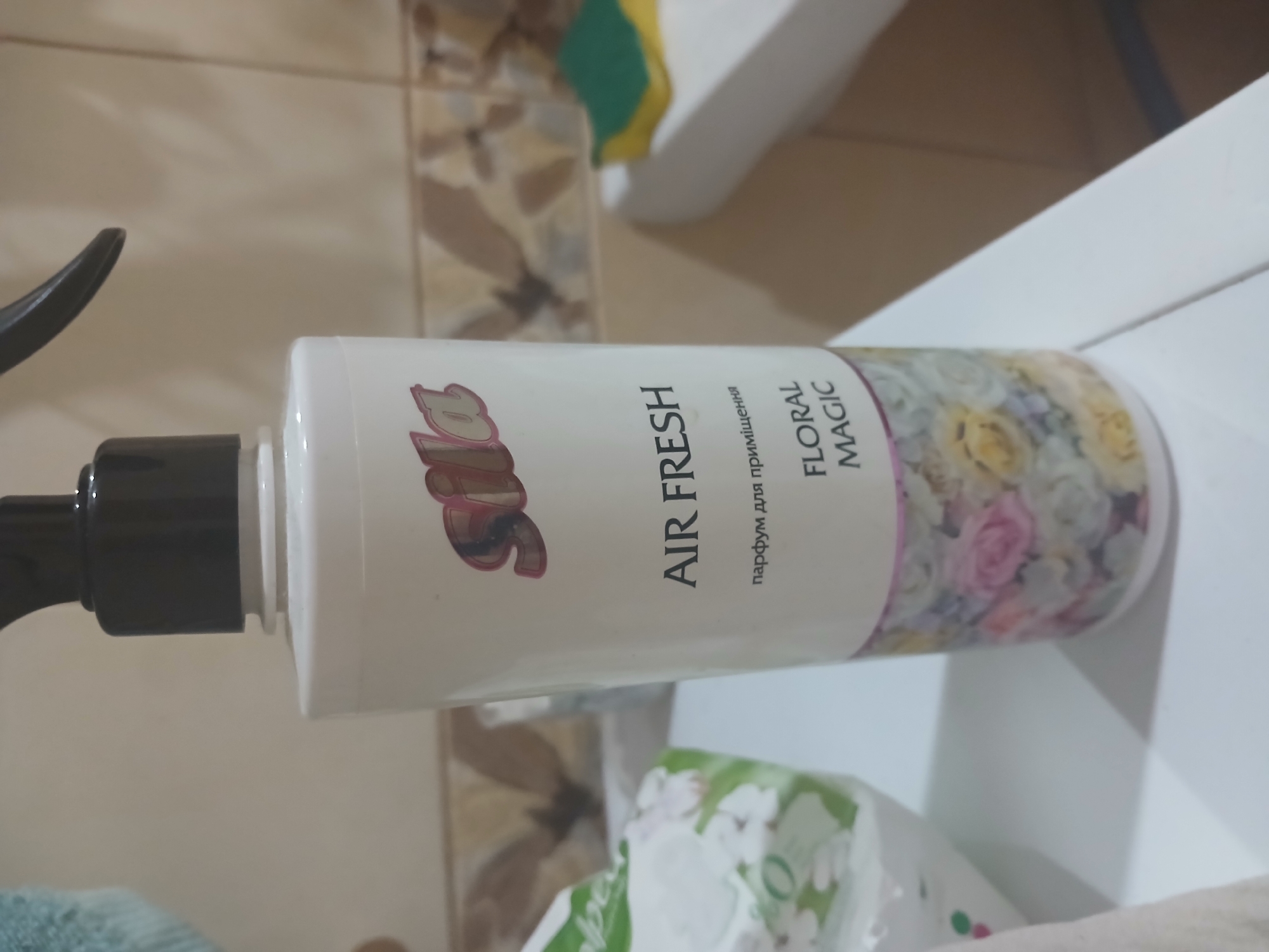 This image shows a bottle of "Sila Air Fresh" floral magic air freshener. The bottle is white with a black spray nozzle. The label features the "Sila" logo, the text "AIR FRESH," and "floral magic." Below "AIR FRESH" in smaller text, it reads "парфум для приміщень" which translates to "perfume for premises." The lower part of the bottle has a floral design.
The bottle is placed on a white surface, possibly a countertop or shelf. To the left, there is a partially visible white package with green accents and floral patterns, suggesting it might be a tissue box or another household product. In the background, a tiled wall with neutral-toned tiles is visible, and to the right, a white bathtub with a yellow and green sponge is present. The overall environment appears to be a bathroom or a similar domestic setting.
The image does not provide any information about the city of Grigoriopol, Moldova.
