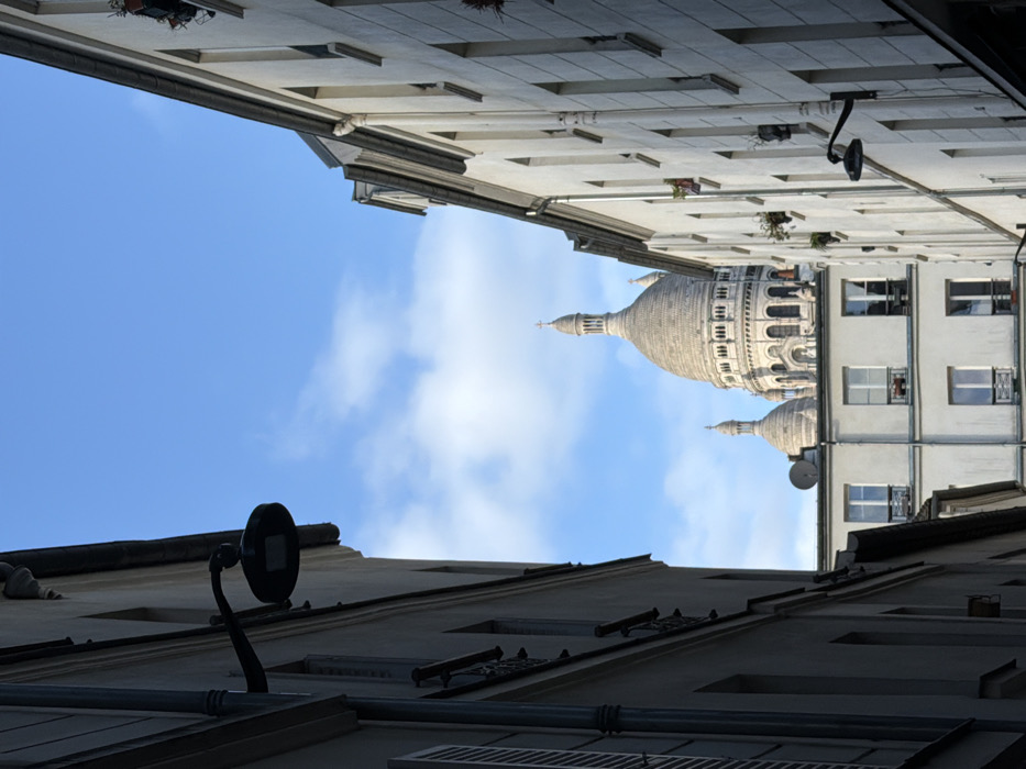 The image depicts a view looking upwards in a Parisian alley. The architecture is characterized by tall, white buildings with symmetrical windows and wrought-iron details. The Sacré-Cœur Basilica's domes punctuate the skyline, framed by a blue sky with scattered clouds. Utility lights are attached to the buildings, adding a contemporary element to the classic European scene.