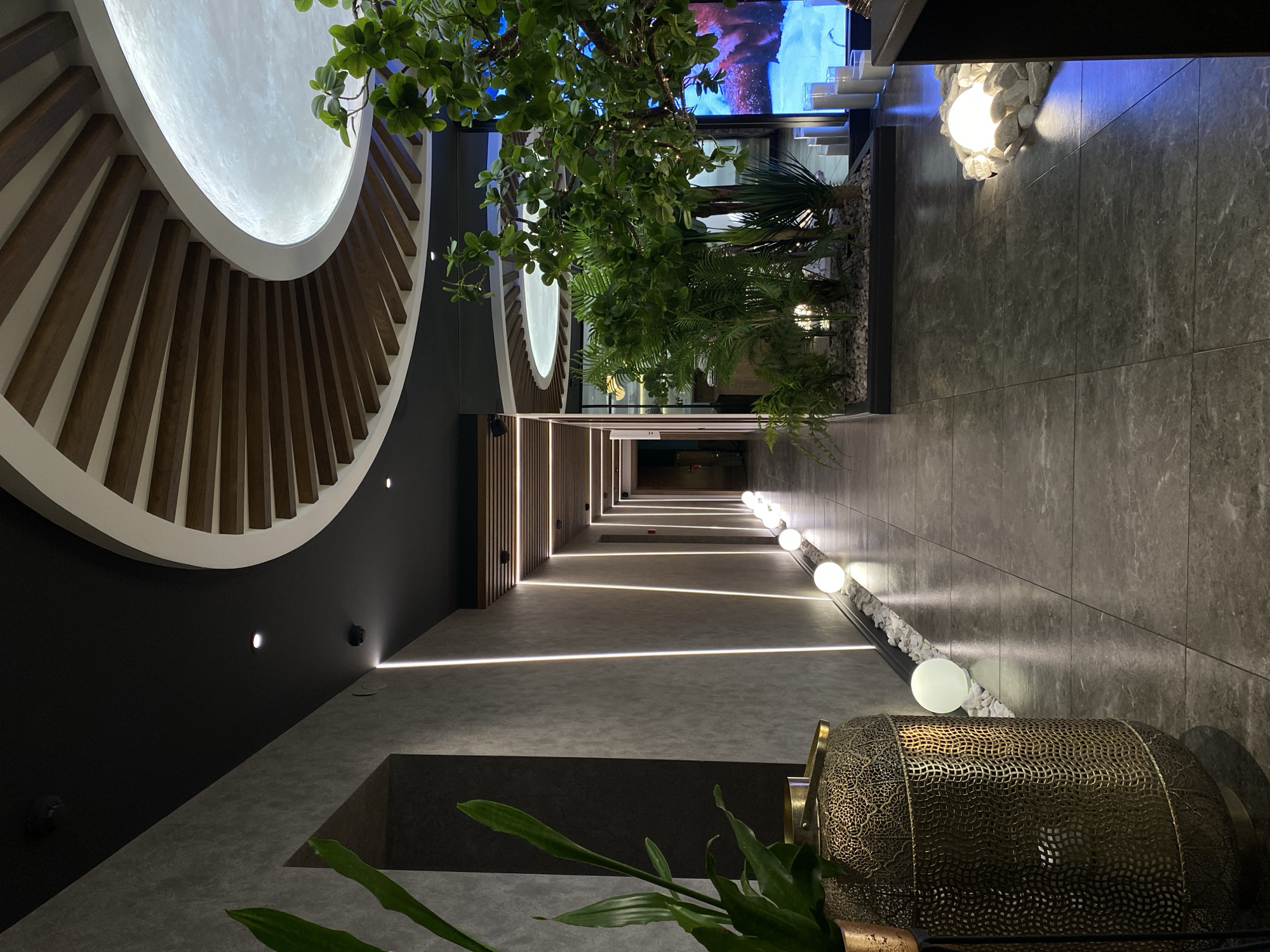 The image depicts a hallway interior. A large, illuminated circular design element is on the left of the ceiling. A line of foliage extends along the hallway. Linear lights extend down the hallway, leading to a series of spherical lights on the right. A large, cylindrical object with a patterned surface is in the foreground. The location is Votkinsk, Russia. No people or interactions are visible.