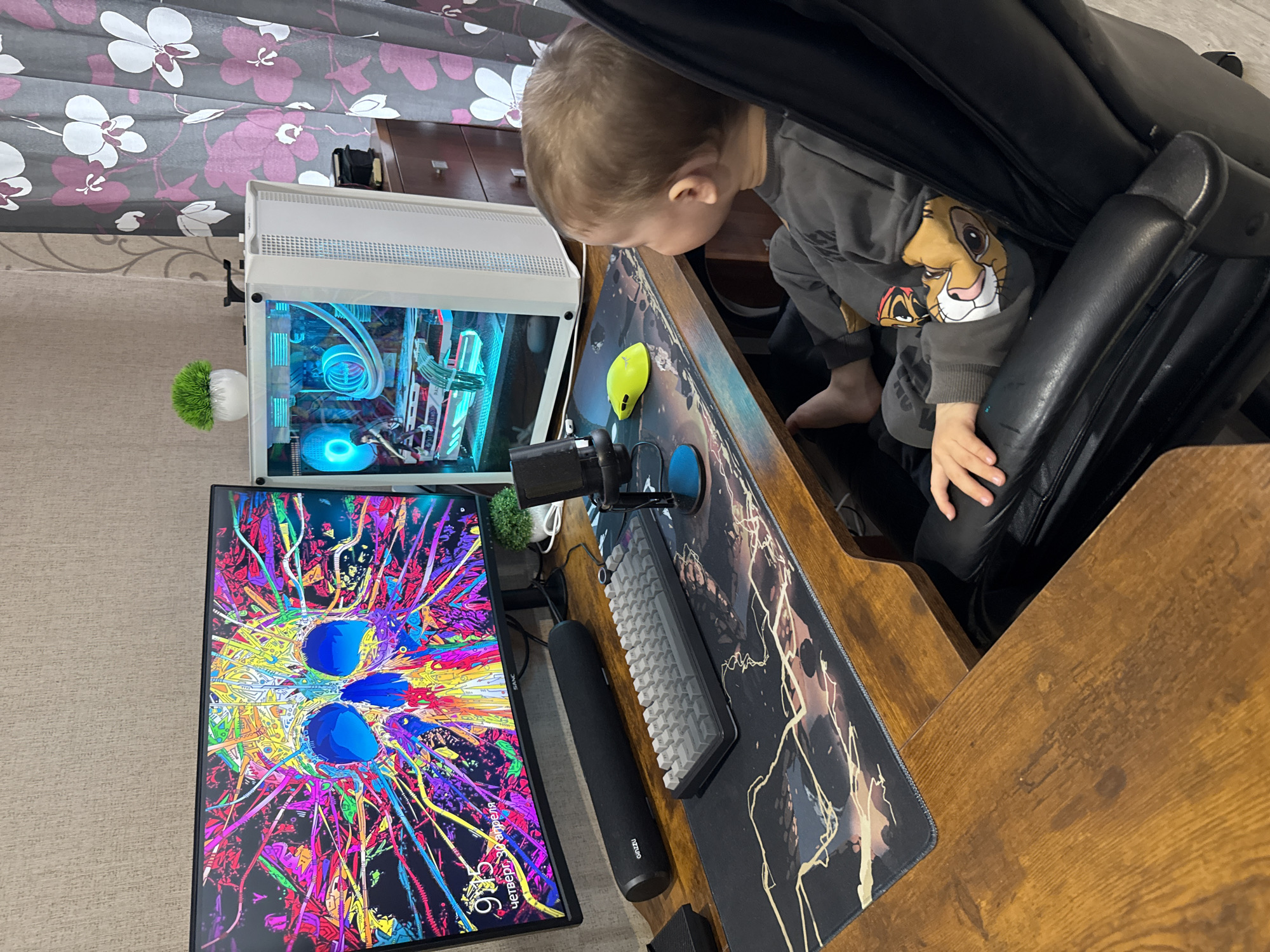 An indoor scene, likely in Rusanovo, Russia, captures a young child's curiosity about a gaming computer setup. A toddler, with light brown hair, is seen leaning forward from a black gaming chair, intently looking down towards a wooden desk. They are wearing a grey long-sleeved top featuring the character Simba from The Lion King, and their bare feet are visible.
The desk holds a high-end PC setup. To the left, a white computer case with a transparent side panel displays vibrant blue and cyan RGB lighting from its internal components, including intricate liquid cooling tubes. Faint text reading "GAMING" is visible inside the case. Adjacent to the PC, a monitor stands in vertical orientation, showcasing a vivid, abstract digital artwork filled with bright colors and two prominent blue circular shapes. On the bottom left of the monitor, partially obscured Russian text indicates "Четверг" (Thursday) and "9:06". The monitor's brand label, beginning with "VENG...", is also visible.
On the wooden desk, a large black mousepad with a striking white and gold abstract design covers the surface. A yellow gaming mouse and a grey mechanical keyboard are positioned on it. A black cylindrical speaker, branded "CREATIVE", rests between the monitor and the keyboard. A microphone on a stand is also part of the setup. A small, round potted plant with green, fuzzy foliage sits atop the PC case.
In the background, grey curtains adorned with a pattern of white and purple flowers are visible, along with a glimpse of dark wooden furniture. The floor appears to be carpeted or covered with a light-colored rug. The image captures a moment of childish exploration within a modern home environment.