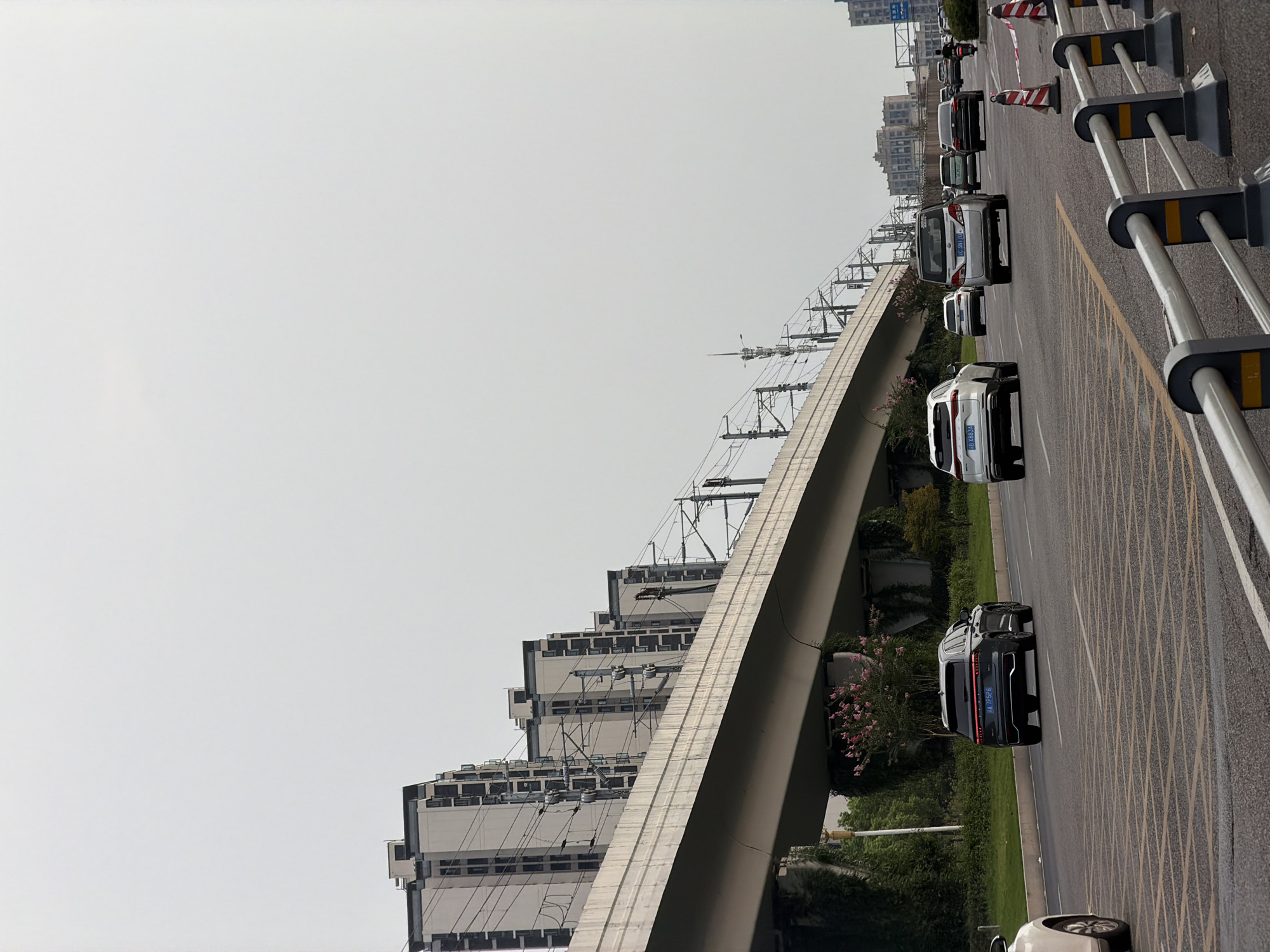 A wide-angle, eye-level outdoor shot depicts a traffic scene on a multi-lane road in Jiangjin, China. The sky is overcast and pale gray. In the foreground, a silver car is visible on the left, partially cropped out. Several other cars, including a dark gray SUV, a white SUV, a silver minivan, and a black sedan, are driving on the road. A motorcycle with a rider is also present. The road has clear lane markings and yellow dashed lines. Metal railings and traffic cones are positioned along the side of the road. An elevated concrete structure, likely an overpass or railway line, runs diagonally across the middle ground, supported by large concrete pillars. Numerous electrical wires and poles are suspended from this structure. In the background, several modern residential buildings with multiple stories are lined up. The overall scene suggests a typical urban environment with ongoing traffic and infrastructure.