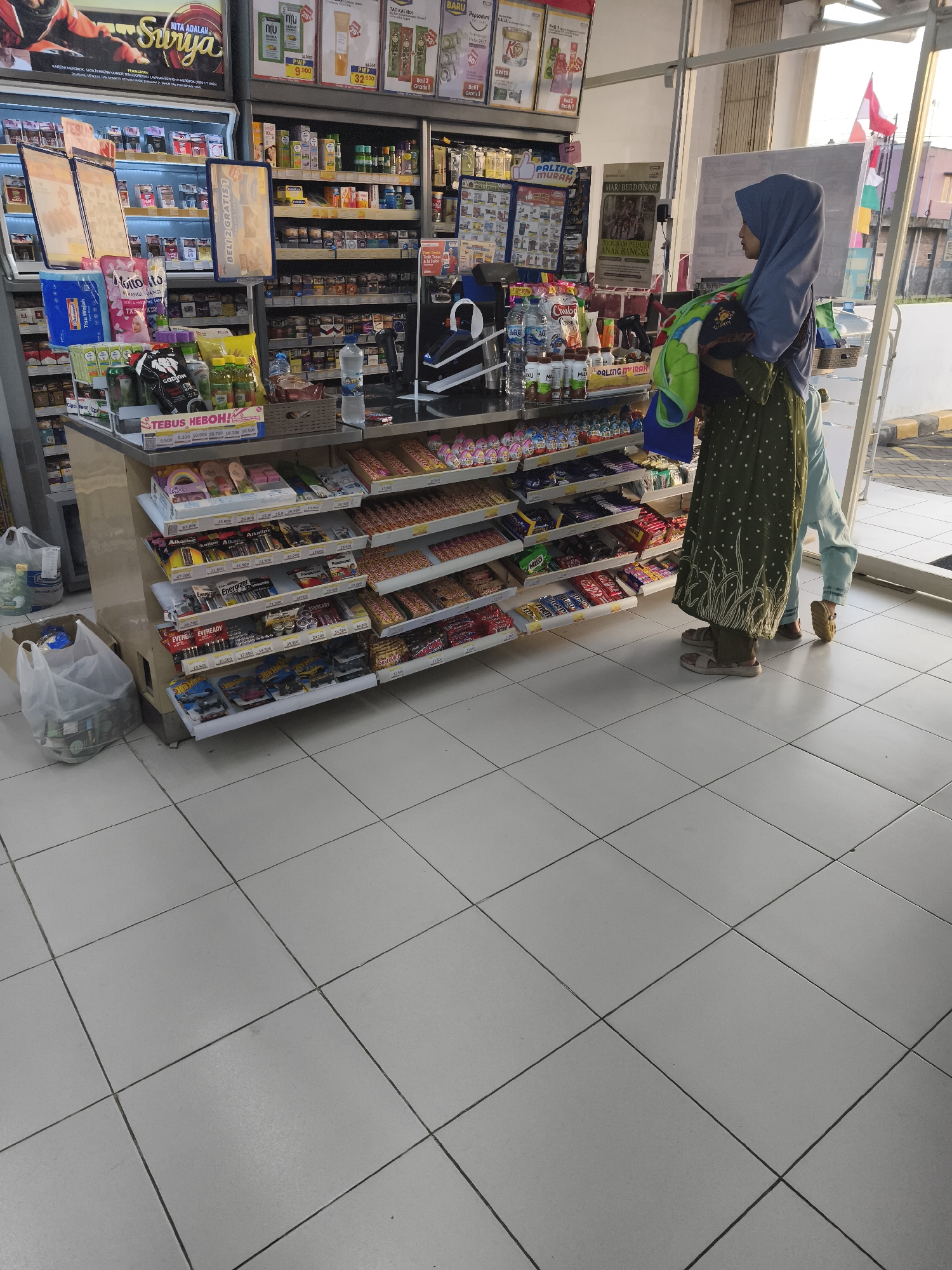 The image shows the interior of a convenience store in Kemiri Kidul, Indonesia. A woman wearing a hijab and a patterned dress is standing in front of a display of snacks, holding a baby wrapped in a blanket. Another person, partially visible, is also in the store. The shelves are stocked with various food items, drinks, and personal care products. The floor is tiled. The overall scene depicts a typical shopping environment in a local Indonesian store.