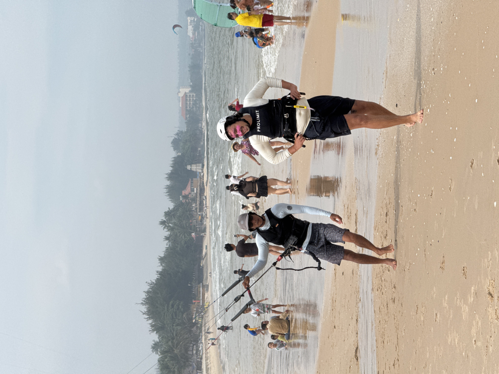 The image captures a lively scene on a beach in Phan Thiết, Vietnam. Two men are prominently featured in the foreground, both engaged in activities related to kitesurfing. The man on the left, wearing a light blue long-sleeved shirt and patterned shorts, is holding kiteboarding control bar and lines. He has a grey baseball cap and sunglasses on. The man on the right, in a white long-sleeved shirt and black shorts, is wearing a white helmet and a black life vest with "PROLIMIT" printed on it. He has pink face paint or makeup on his cheeks and is smiling, with his hands on his hips.
In the background, the beach is populated with numerous other people enjoying the water and sand. Children are playing in the shallow water, and adults are scattered across the beach, some sitting, others walking. The sea is moderately wavy, and in the distance, several kites can be seen in the sky, indicating kitesurfing activity. Palm trees and buildings line the shore, suggesting a developed coastal area. The overall atmosphere is one of a busy, recreational beach day.