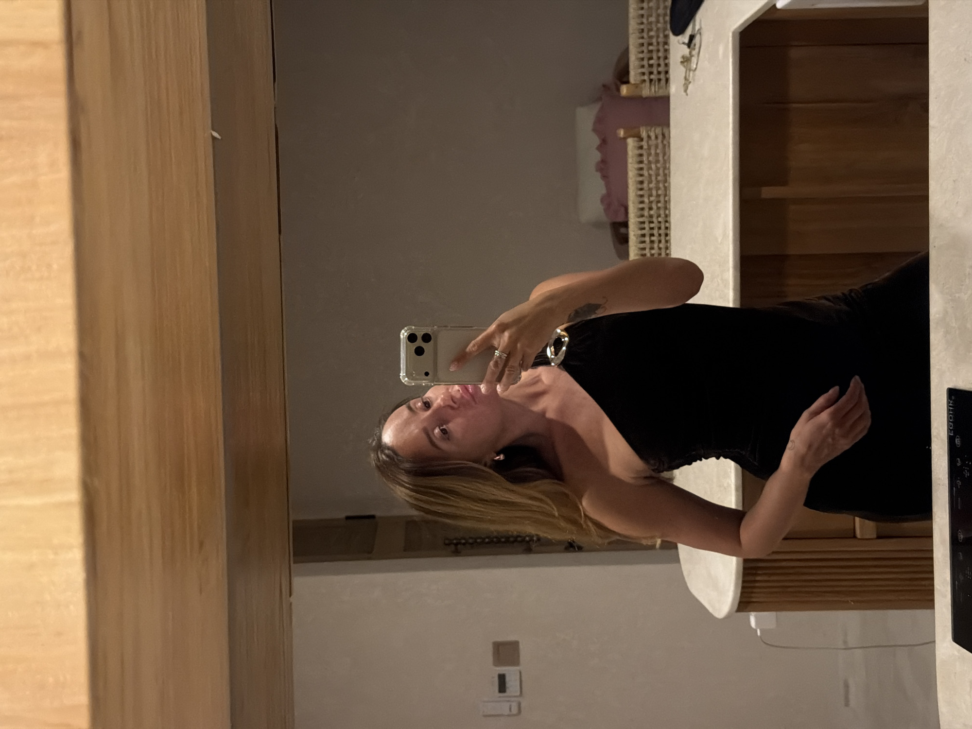 A woman takes a selfie in a mirror, her arm extended to hold her phone. She is wearing a black dress and has blonde hair. She is wearing a ring on her finger. The background shows a room with wooden furniture and a light-colored wall. There is a woven basket visible in the reflection. The scene appears to be indoors.