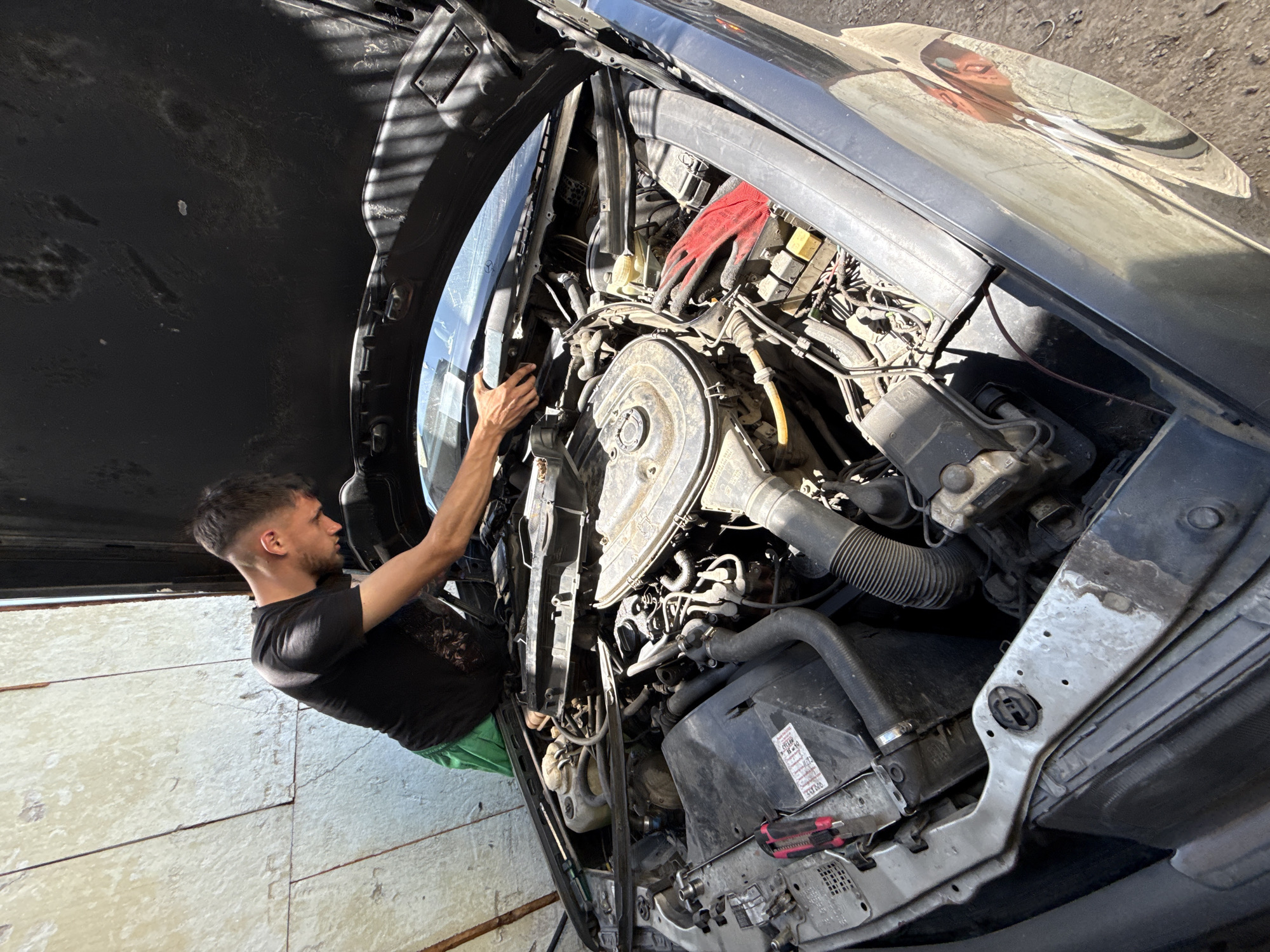 A man wearing a black t-shirt and green shorts is shown working on the engine of a car with its hood open. The car's engine compartment is visible, revealing various mechanical components and a pair of red gloves. The man is positioned on the left side of the frame, reaching into the engine bay. The car's exterior is visible on the right side of the frame. The scene appears to be indoors, possibly in a garage or workshop, with a wall visible in the background. Based on the location of the image, this is in Shumen, Bulgaria.