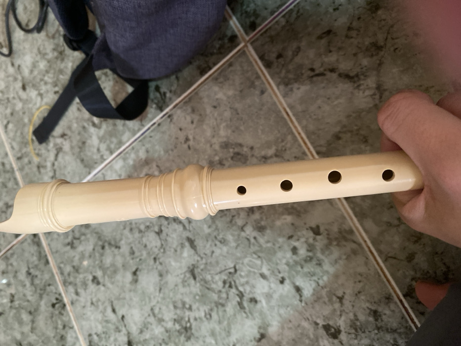 The scene depicts a light-colored recorder held by a person's right hand, positioned vertically against a tiled floor in Đak Đoa, Vietnam. The recorder, appearing cream or light beige, features a visible mouthpiece section, a middle section with two decorative ribbed bands, and a lower section displaying four open circular finger holes. The hand grasps the lower part of the instrument. The floor consists of square, light gray tiles with darker gray patterns, separated by light brown grout lines. In the upper right background, a dark gray fabric bag or backpack is visible, accompanied by a black strap and a thin yellow string or cable. A reflective silver metal rod or pipe extends diagonally across the floor from the upper right to the lower left, passing behind the recorder and the bag. A partial view of a foot or leg, possibly clad in dark fabric, is present in the lower left corner of the frame.