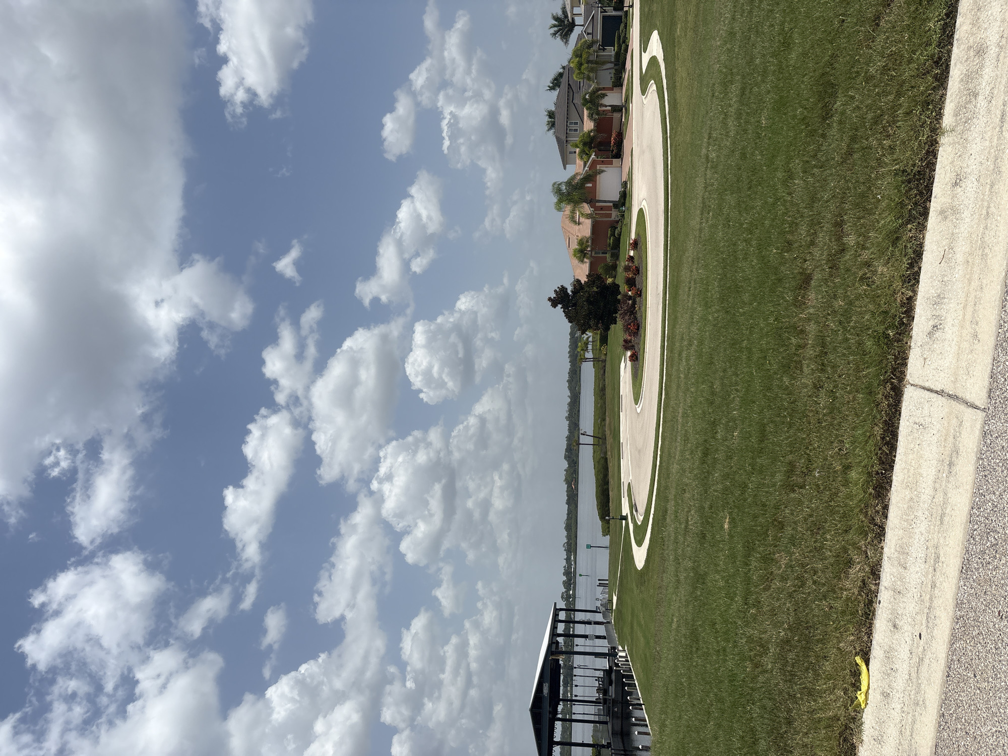 The image, captured in Bradenton, USA, depicts a waterside residential area under a partially cloudy sky. The scene is oriented vertically, requiring a 90-degree rotation for proper viewing. In the foreground, a neatly manicured green lawn slopes gently towards a body of water. A paved pathway with a white border curves through the lawn.
Along the water's edge, a dark, modern boat dock with a canopy structure is visible. The water itself is calm and reflects the sky. Across the water, a line of trees forms a natural border, with a few houses peeking through the foliage. These houses appear to be modern in design, with tiled roofs.
The sky is a vibrant blue, dotted with fluffy white cumulus clouds, suggesting fair weather. The lighting indicates it is likely daytime, possibly late morning or early afternoon. The overall atmosphere is serene and suggests a peaceful, affluent neighborhood. There are no people or specific activities clearly depicted, focusing instead on the landscape and architecture. No visible text is present in the image.