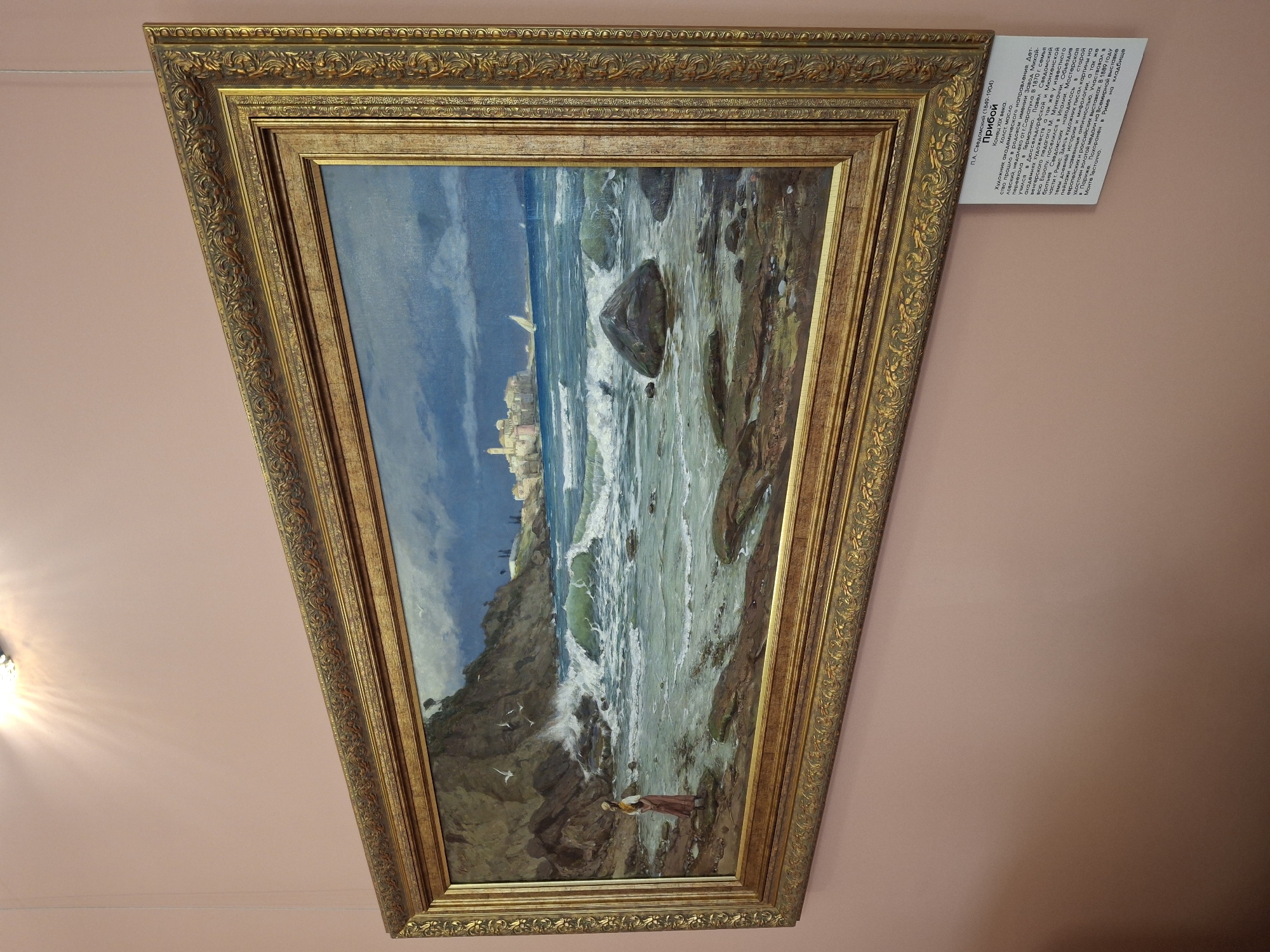 A painting titled "Прибой" (Priboy, meaning "Surf" or "Breaker") by P.A. Svedomsky (Павел Александрович Сведомский, 1849-1904) is displayed on a light pink wall. The artwork, from the late 19th century and executed in oil on canvas, is housed in an ornate, gilded wooden frame.
The painting depicts a vivid seascape with a rugged, rocky coastline in the foreground. Powerful, foamy waves crash against the shore, creating a dynamic and turbulent scene. The sea transitions from a deep blue to a lighter, frothy green where the waves break. In the mid-distance, a small, light-colored town or village with distinct buildings is situated on a hill overlooking the water. A sailboat is visible on the distant horizon near the town. The sky is a clear blue, suggesting daylight.
In the bottom right foreground, a solitary figure, likely a woman dressed in a long, dark garment and a light-colored head covering, stands on the rocks, gazing out at the sea. A few white birds, possibly seagulls, are depicted flying low over the rocks on the left side.
Adjacent to the painting, a white information panel provides details in Russian. It identifies the artist, title, and states the painting was created in the "Конец XIX века" (End of the 19th century) using "Холст, масло" (Canvas, oil). The text also includes a biography of Svedomsky, mentioning his academic training, studies in Germany (Düsseldorf and Munich), his relocation to Italy (Rome) where he focused on ancient history, mythology, and landscapes, and his participation in exhibitions including a gold medal at the 1889 World Exhibition in Paris. The artist's childhood was spent in Sarapul, Russia, before his family moved to Germany. The panel also notes his burial in Rome.
The wall on which the painting is displayed is a soft pink, and a light fixture, possibly a chandelier, is partially visible in the top-left corner of the frame, casting bright rays.