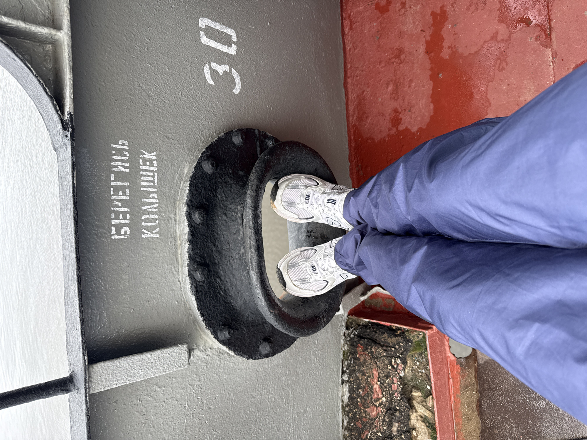 A person's legs, clad in blue trousers and white athletic shoes, are positioned inside a large, circular opening on the side of a grey metallic structure. The opening is reinforced with a thick, black rubber gasket and secured with numerous bolts. White Russian text is stenciled on the grey surface above the opening, reading "БЕРЕГИСЬ КОЛЬШЕК" (Beware of bumps). To the right of the opening, the number "30" is stenciled in white. The structure appears to be part of a boat or ship, with a red, wet deck visible below. The background shows the surface of the water. The scene is likely located in the city of Kozmodemyansk, Russia, given the Russian text.