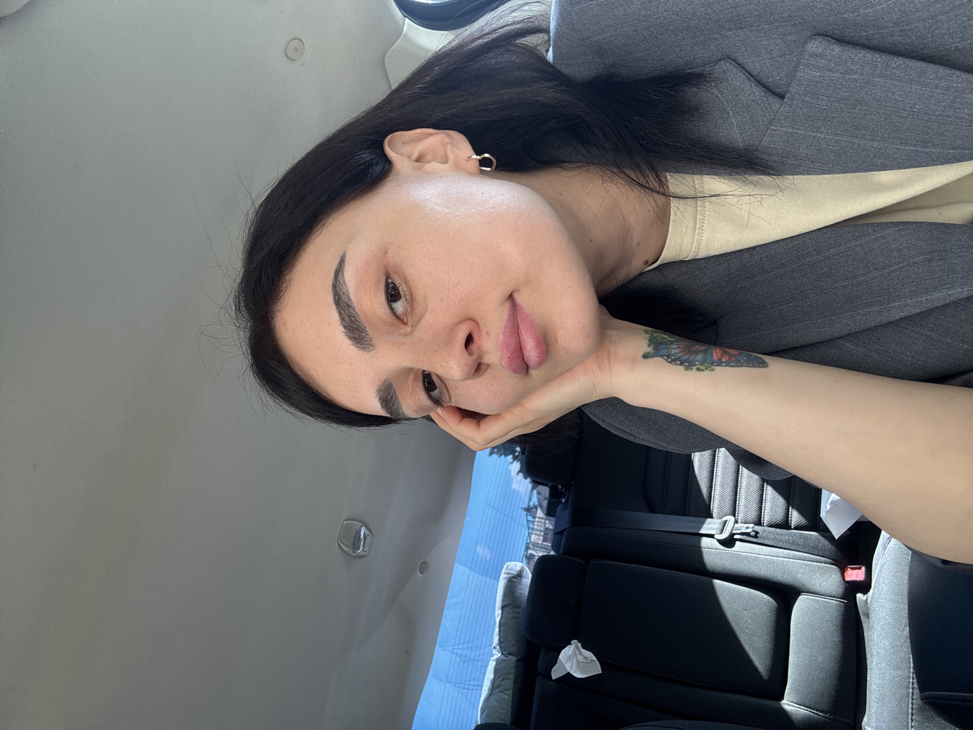 The image depicts a woman inside a car. She has dark hair, prominent eyebrows, and a butterfly tattoo on her forearm. She is wearing a beige shirt and a gray blazer. The car's interior features black leather seats. The background shows a glimpse of the outside, including a building. Based on visual cues, the location is potentially in Tbilisi, Georgia.