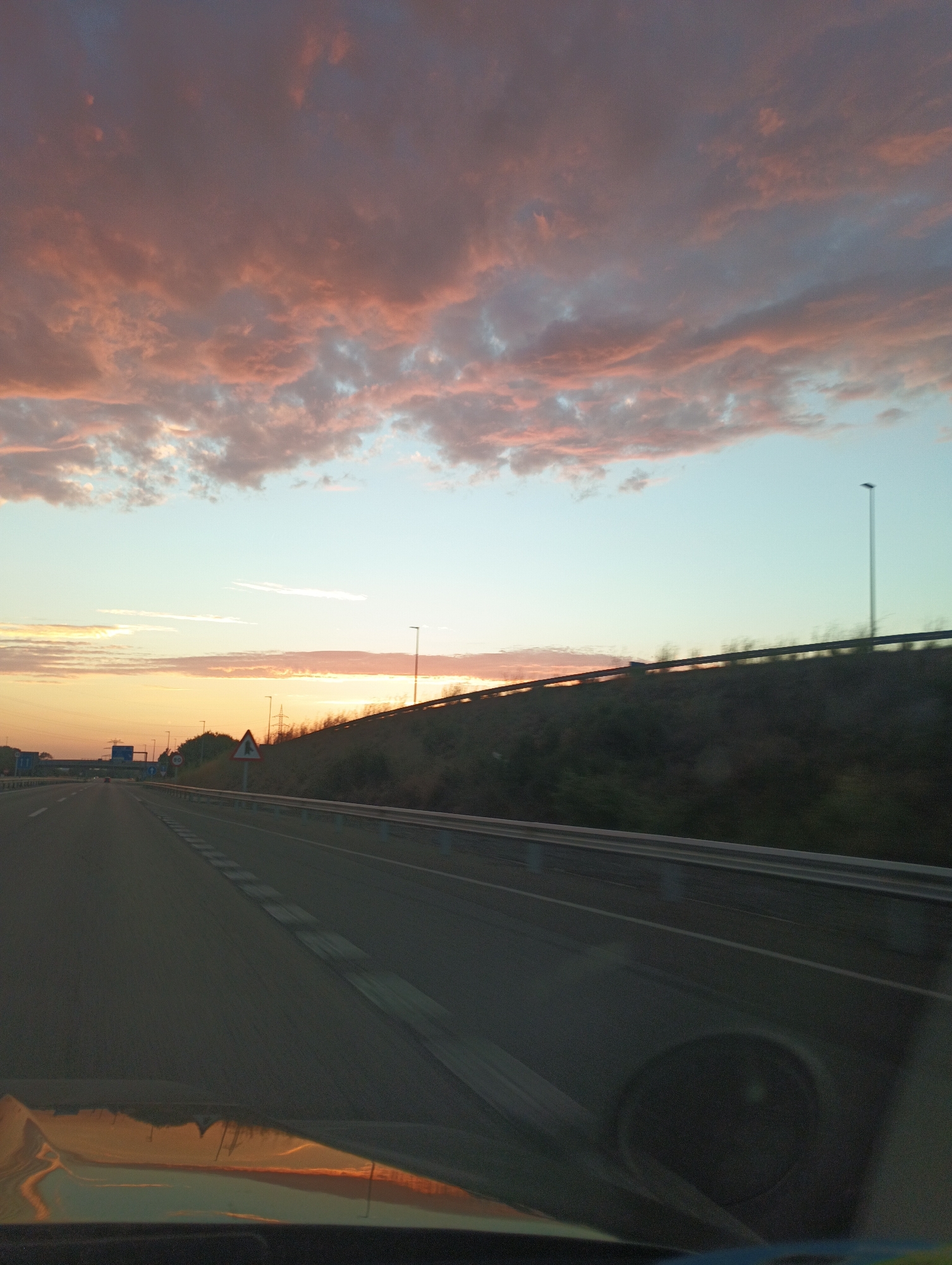 The image depicts a highway scene at sunset. The sky is filled with pink and orange clouds, illuminated by the setting sun. A road stretches into the distance, with guardrails and lighting poles lining the side. The perspective suggests the photo was taken from inside a vehicle. Vegetation is visible on the embankment beside the highway. The overall impression is one of a tranquil, scenic drive as day transitions to night.