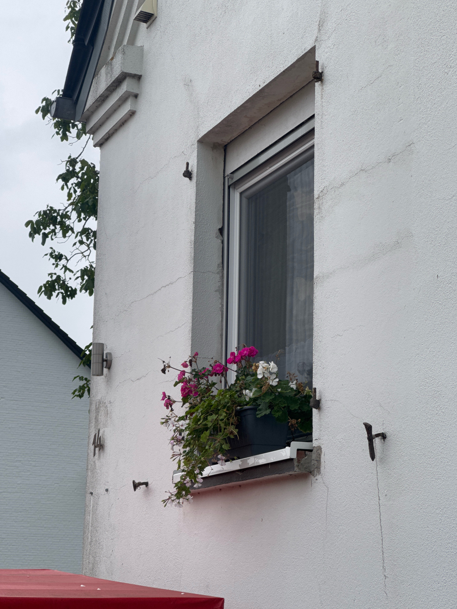 The image captures a close-up view of a white stucco building facade in Cleves, Germany, featuring a window box filled with pink and white flowers. A dark grey gutter system is visible along the roofline on the left side of the frame, partially obscured by the branches of a leafy green tree. To the left of the building, a portion of a neighboring white brick building with a dark gabled roof is visible. On the white facade, several dark, rusty metal fixtures are attached at varying heights, including what appears to be a number "14" at mid-level. The window itself is a double-pane unit with a grey frame, reflecting the overcast sky. The window box is supported by a white ledge, and below this, a red awning or covering extends from the bottom edge of the frame. The stucco wall shows signs of age, with fine cracks present.