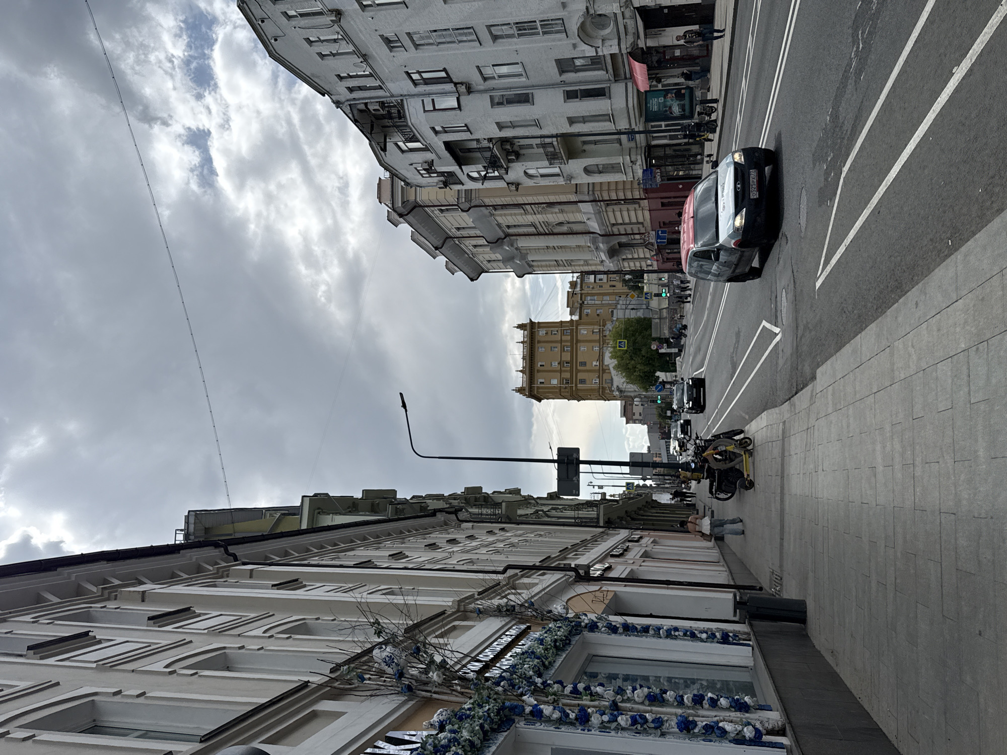 This is an eye-level, full-shot, outdoor, daytime photograph of a street scene in Moscow, Russia. The image features several multi-story buildings lining both sides of a paved street. The buildings on the left are predominantly light-colored with white trim and elaborate architectural details, including columns and decorative elements. One building on the left has blue and white floral decorations near its entrance. The buildings on the right are a mix of colors, including white and beige, and also display ornate facades and balconies. In the distance, a tall, yellow-colored building with a distinct architectural style is visible.
The street is multi-laned with clear white lane markings. A light-colored van with a red roof is driving on the right side of the road. Several other vehicles are also present on the street. On the sidewalk to the left, a few people are walking. Two electric scooters are parked on the sidewalk. A lamppost stands on the sidewalk in the foreground. The sky is overcast with heavy, gray clouds, suggesting a dull or possibly rainy day. The overall atmosphere is urban and busy.