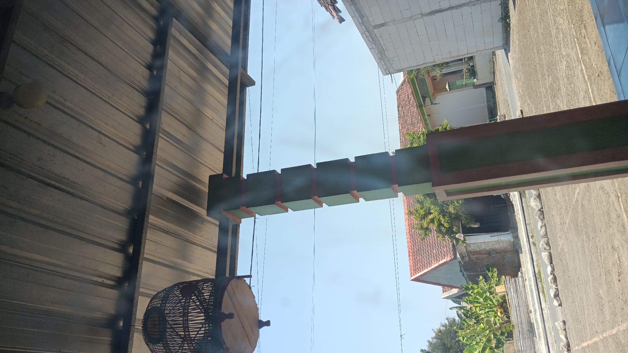 The image depicts an outdoor scene in Sonobekel, Indonesia. In the foreground, a tall, green, and brown rectangular pillar with segmented sections dominates the right side of the frame. To the left and above, a birdcage made of woven material hangs from the corrugated metal roof of an overhang. Behind the pillar and birdcage, a clear blue sky serves as the backdrop. Further in the background, several buildings are visible: a house with a red-tiled roof and grey brick walls, another building with a white brick facade, and a smaller structure with a red-tiled roof and brick walls. A banana tree with large leaves is situated to the left of the buildings. The ground in front of the buildings is a dirt or gravel surface, with a line of rocks forming a border. The overall impression is that of a residential or semi-rural area.