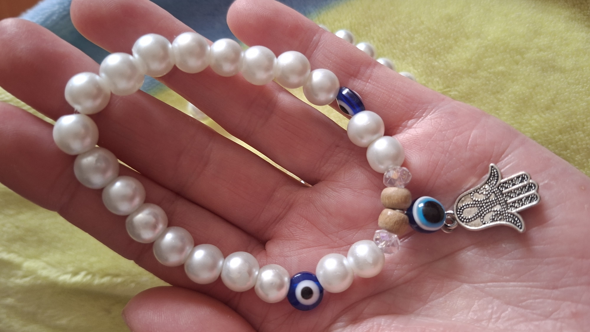 A hand is holding a beaded bracelet in Забалоцкі сельскі Савет, Belarus. The bracelet consists of white pearl-like beads interspersed with a dark blue evil eye bead, wood beads, and clear faceted beads. A silver-colored Hamsa charm is attached to the bracelet. The background is a soft, yellow fabric.