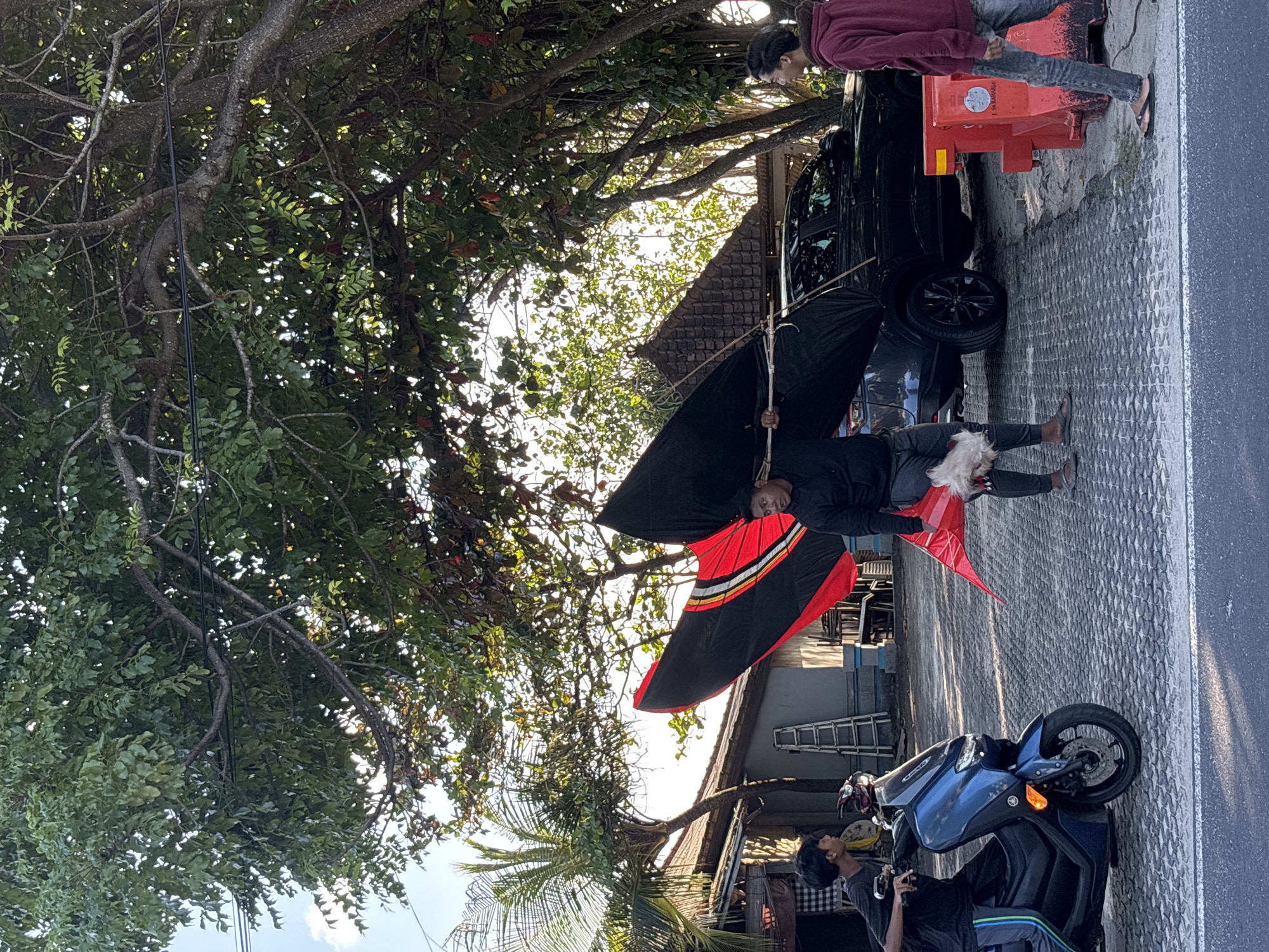 The image shows a daytime scene in Kuta, Indonesia. A person in dark clothing stands in the street holding a large kite with red, black, and white coloring. Another person stands nearby on the sidewalk. A black car and a blue motorcycle are also present in the background, alongside a building with a tiled roof. A large tree with dense foliage partially obscures the sky.