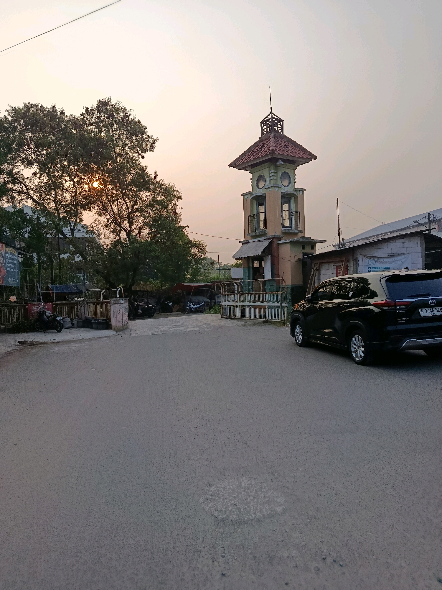 The image shows a street scene in Bekasi, Indonesia, with a distinctive tower and a black SUV. The time of day appears to be late afternoon or early evening, with the sun setting behind a large tree on the left side of the frame. The sky is a soft orange and yellow hue.
In the foreground, a paved road with some scattered debris occupies the lower half of the image. To the right, a modern black SUV is parked facing away from the camera, with its license plate visible: B 1446 HZD. The SUV has tinted windows and appears to be a relatively new model.
Dominating the middle ground on the right is a multi-story structure that resembles a clock tower or a guard post. It is painted in pastel colors, primarily light green and peach, with a red tiled roof and decorative accents. There are windows or openings on its upper levels. Next to the tower, on the far right, is a low-rise building with white brick walls and a corrugated metal roof. A banner or sign is visible on this building, with some text in Indonesian.
To the left of the tower, a large tree with dense foliage partially obscures the setting sun. Underneath and around the tree, there are various parked motorcycles, some makeshift shelters covered with tarps, and what appears to be a metal fence or barrier. A large billboard with colorful graphics is also visible on the far left. The overall impression is a somewhat utilitarian and developing urban environment.