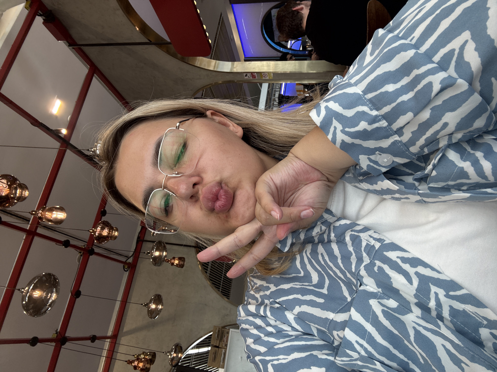 A woman with blonde hair and glasses is taking a selfie in a dimly lit restaurant. She is wearing a blue and white patterned shirt and has her hand on her chin with two fingers extended, making a peace sign. Her lips are pursed, and she is looking at the camera. In the background, there are several hanging lights and arches that suggest an indoor dining setting. The overall ambiance is casual and relaxed.